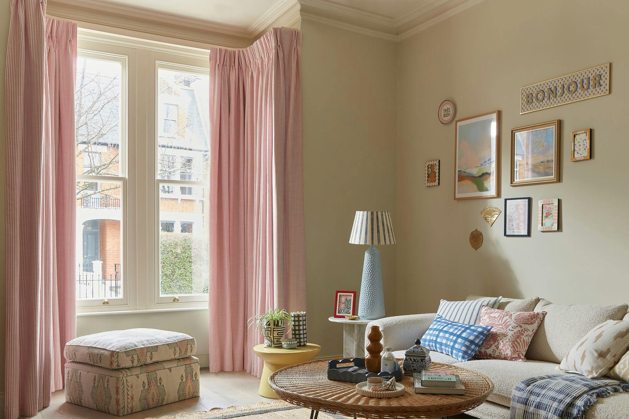 Room features a cozy sofa with colorful pillows, a wicker coffee table with decor, and a side table with a lamp. Pink curtains frame large windows. Wall art includes "BONJOUR" text.