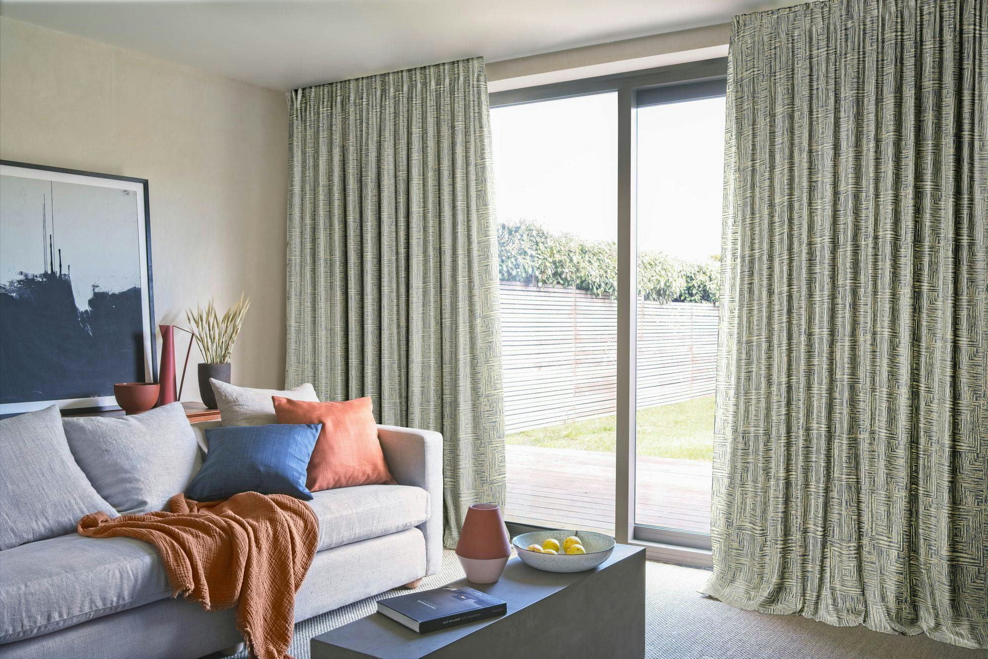 A gray sofa adorned with colorful pillows and a blanket sits in a living room beside a modern coffee table. A large window with patterned curtains overlooks a garden patio.