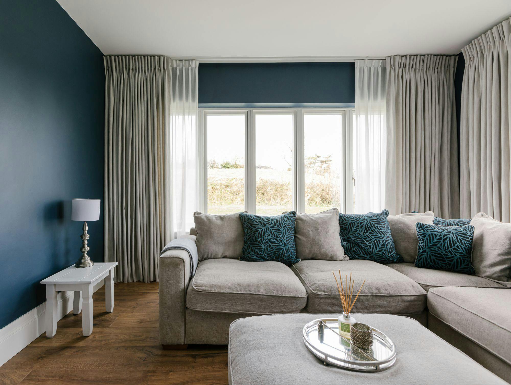 A beige sectional sofa with blue-patterned cushions sits in a living room, accompanied by a small side table with a lamp. Large windows with curtains illuminate the hardwood floor.