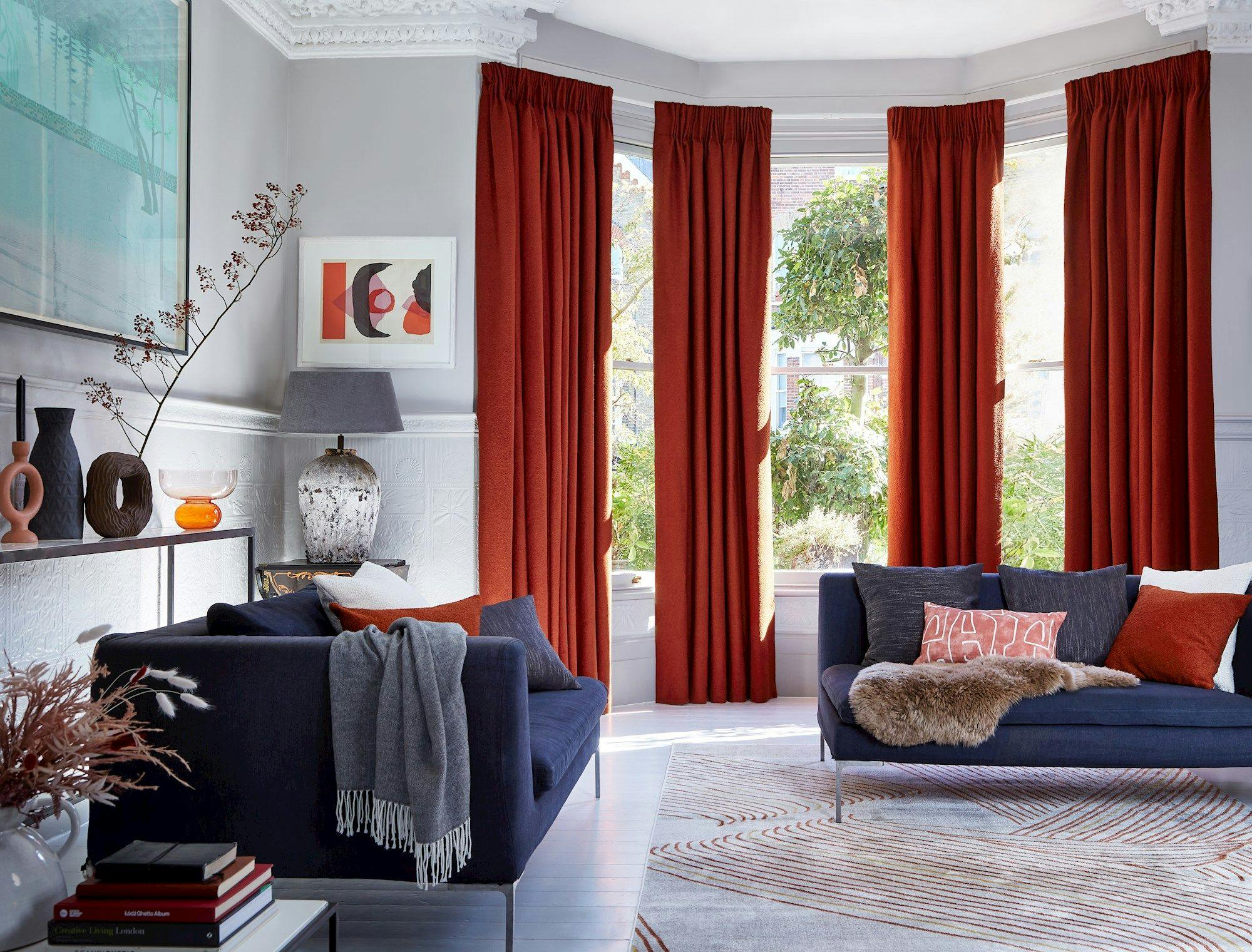 Sofas with colorful cushions sit in a living room. Red curtains frame large windows, allowing natural light. Decorative vases and a lamp rest on a console table. A patterned rug covers the floor.