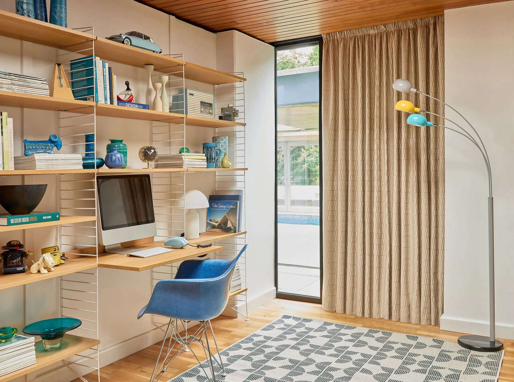 A study area features a computer on a wooden desk, surrounded by books, decorative items, and a blue chair. It is beside a window with beige curtains and a colorful floor lamp.