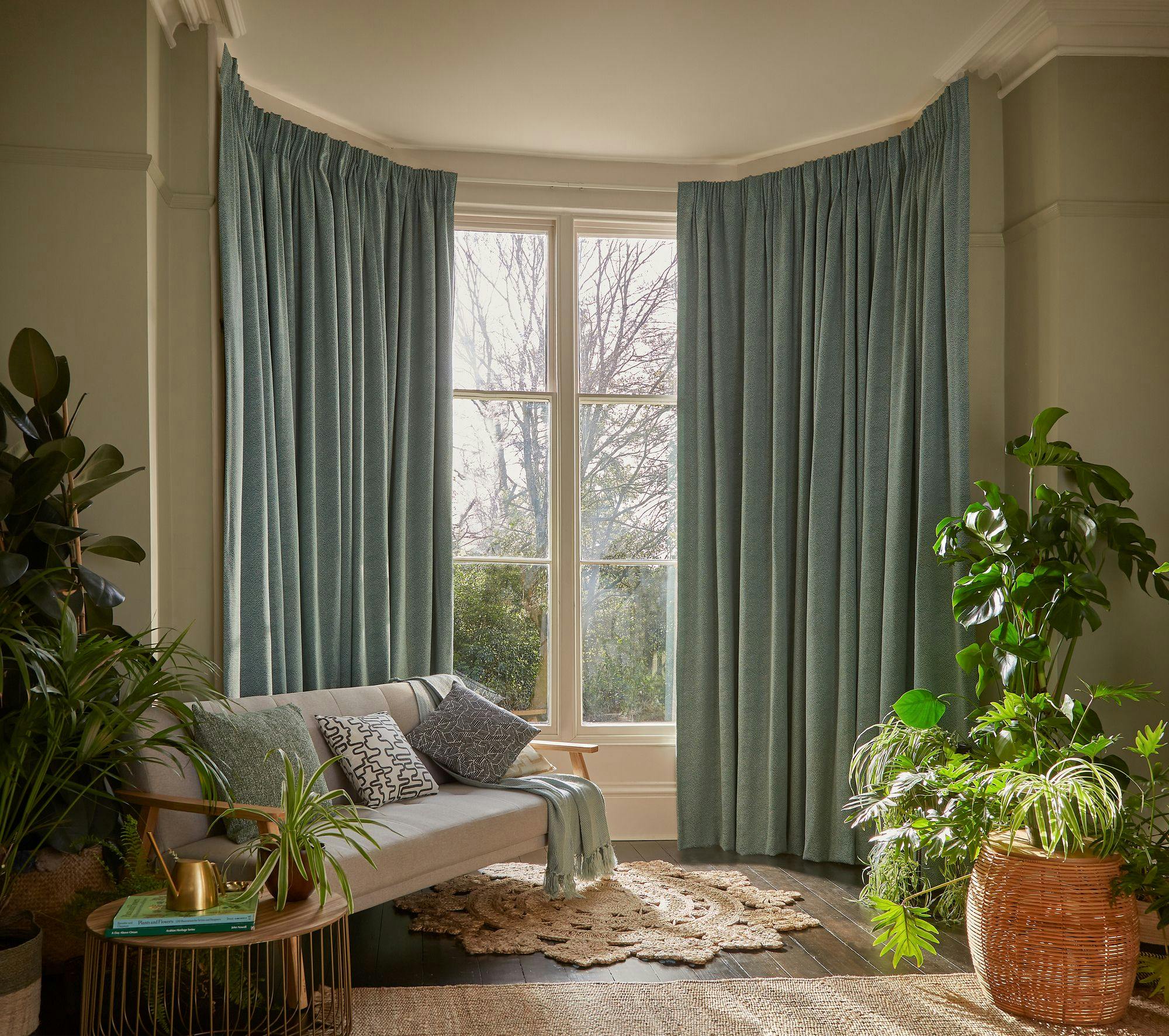 A cozy living space features a light gray sofa with decorative pillows, surrounded by potted plants. Blue curtains cover large windows, allowing soft natural light to enter.