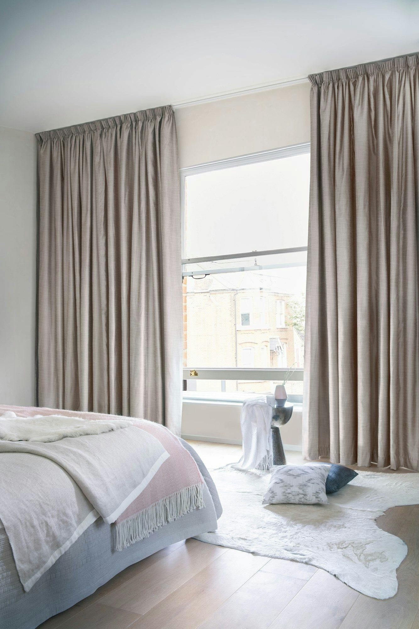 A bed with layered blankets and sheeting sits beside a large window with beige curtains in a bright bedroom. A cozy rug and pillow lie on the wooden floor.