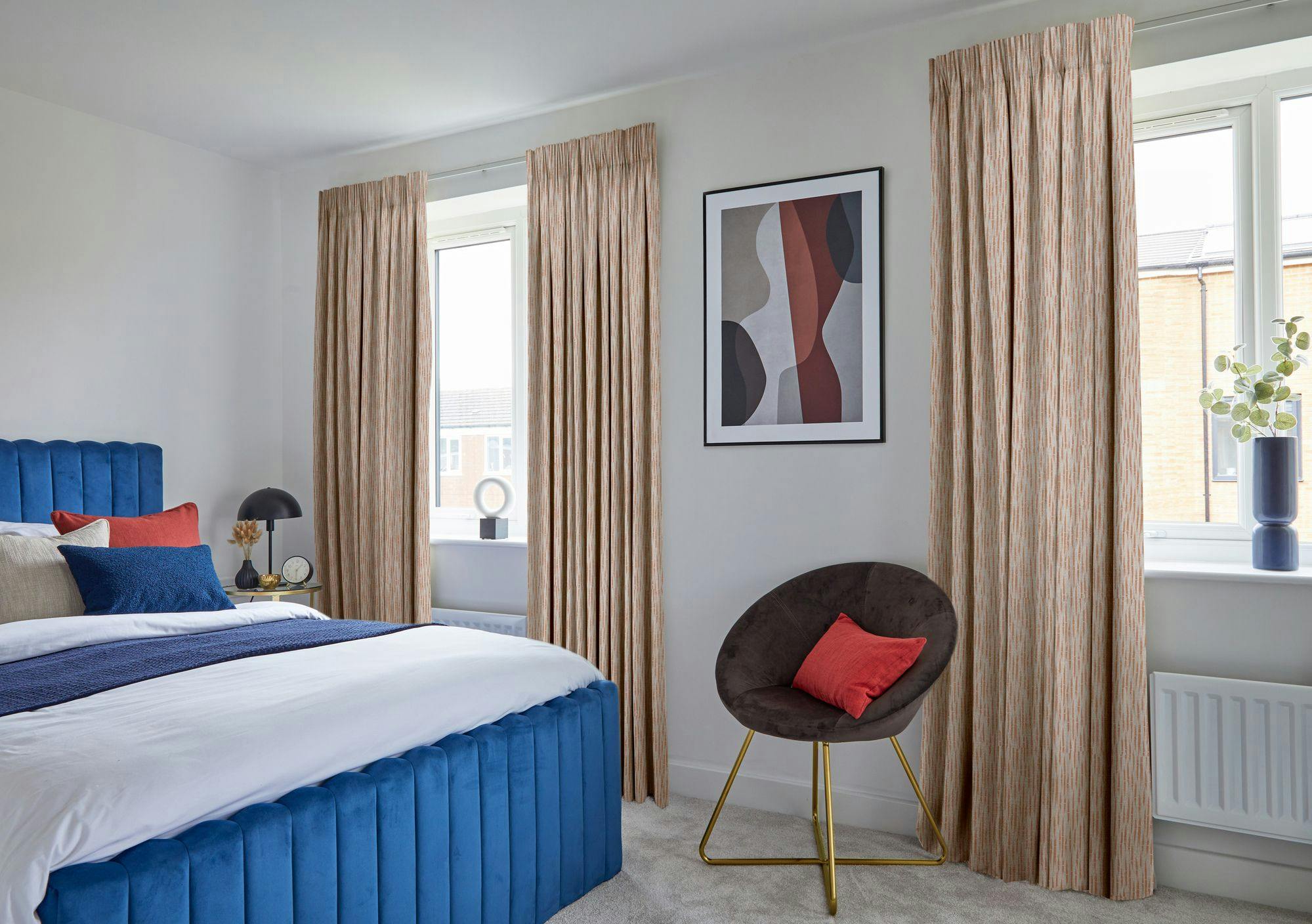 A bed with a blue frame and colorful pillows rests beside a table with a lamp and clock. Two windows with curtains, a chair, and abstract wall art complete the room's decor.
