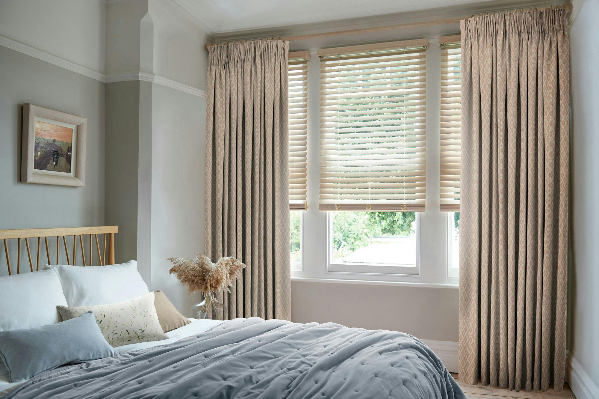 A bed with pillows and a gray comforter is situated in a cozy bedroom, beside a window with beige curtains and blinds. A picture hangs on the pale green wall.