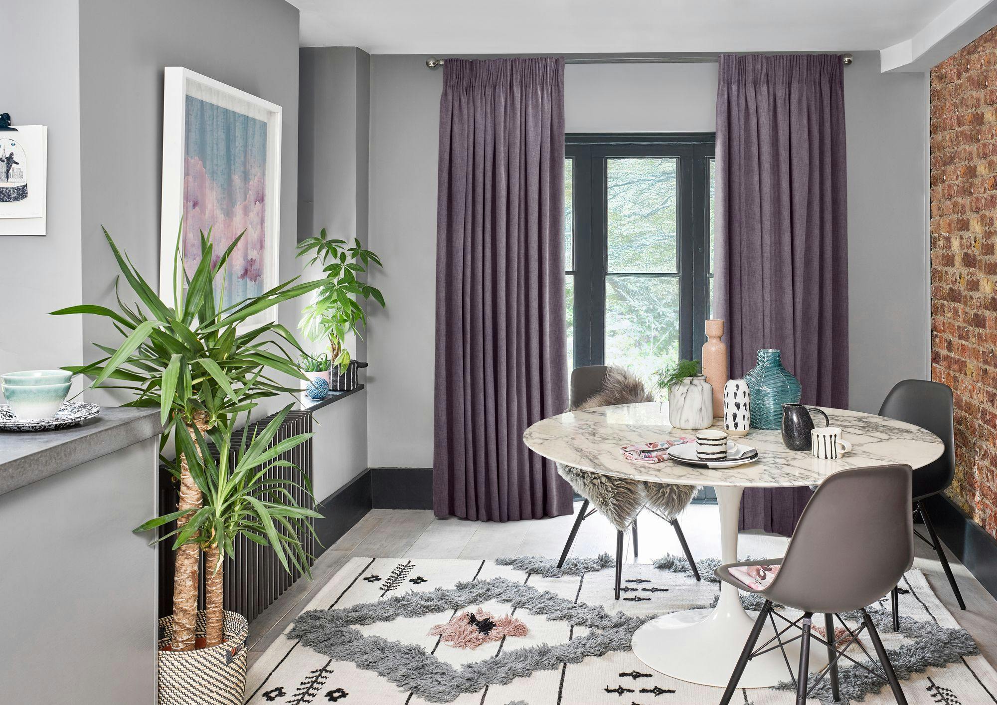 A round marble table with assorted decor and chairs stands in a gray-walled room with a brick accent, purple curtains, large plants, a framed picture, and a patterned rug.