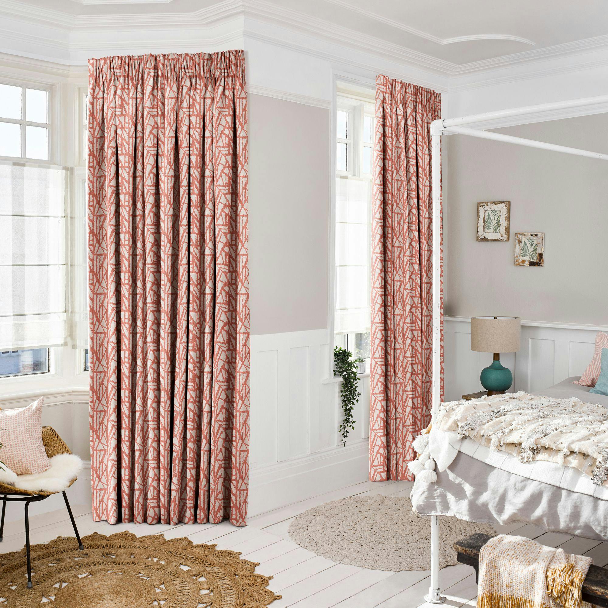 Pink patterned curtains hang beside a window, complementing a cozy bedroom with a white bed, teal lamp, textured rug, and framed botanical prints on a beige wall.