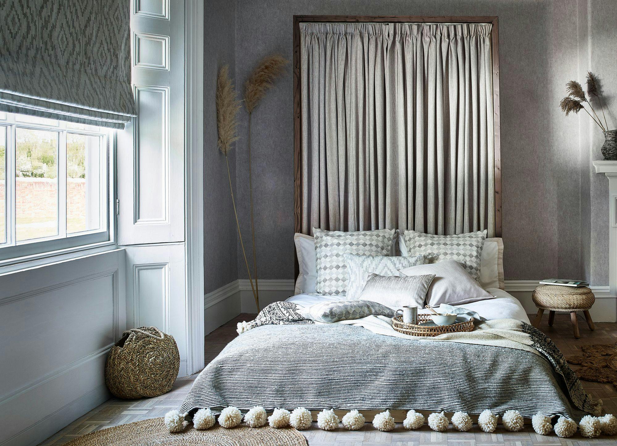 Bed with patterned pillows lies under a textured blanket in a softly lit room. Curtains frame the bed, and pampas grass decorates the corner near a window.
