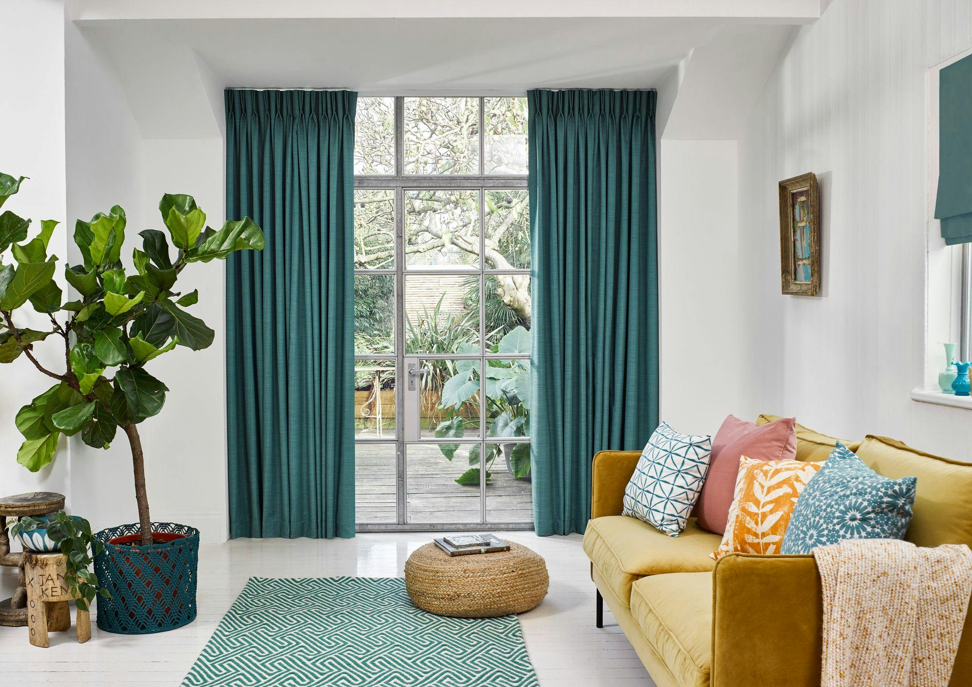 A fiddle leaf fig tree stands beside teal curtains framing a window. A mustard sofa with patterned cushions sits on a geometric rug; a round woven ottoman is nearby. Light floods the room.
