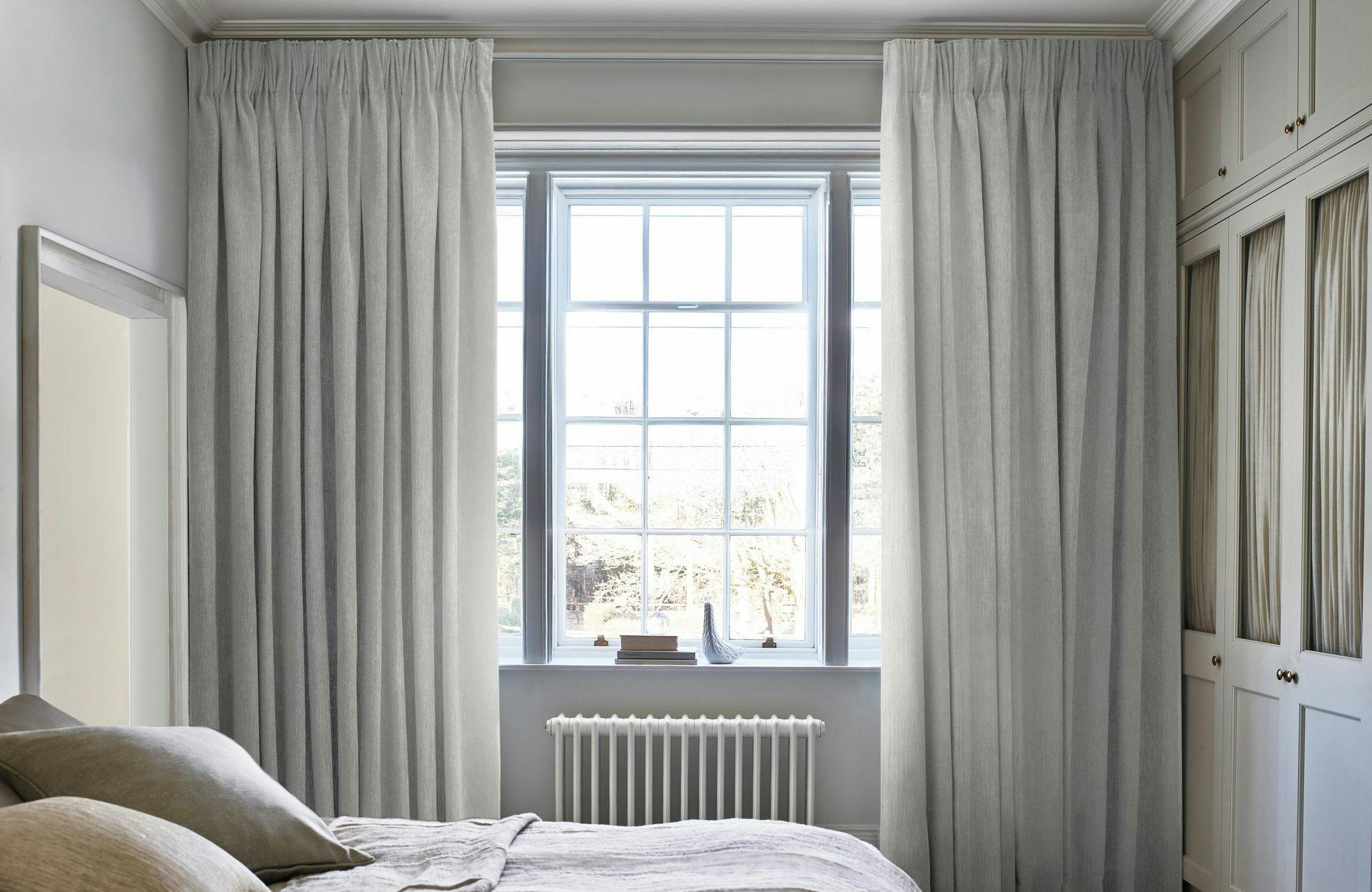 A bed with beige linens rests against a large window with light curtains. A radiator and cabinets are visible in a calm, softly lit bedroom.