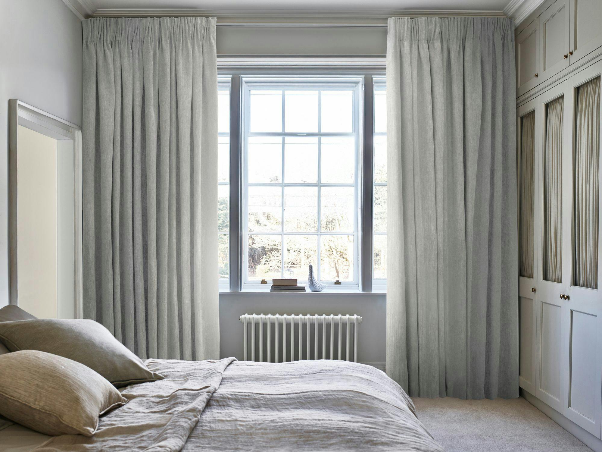 A bed with beige linens rests against a large window with light curtains. A radiator and cabinets are visible in a calm, softly lit bedroom.