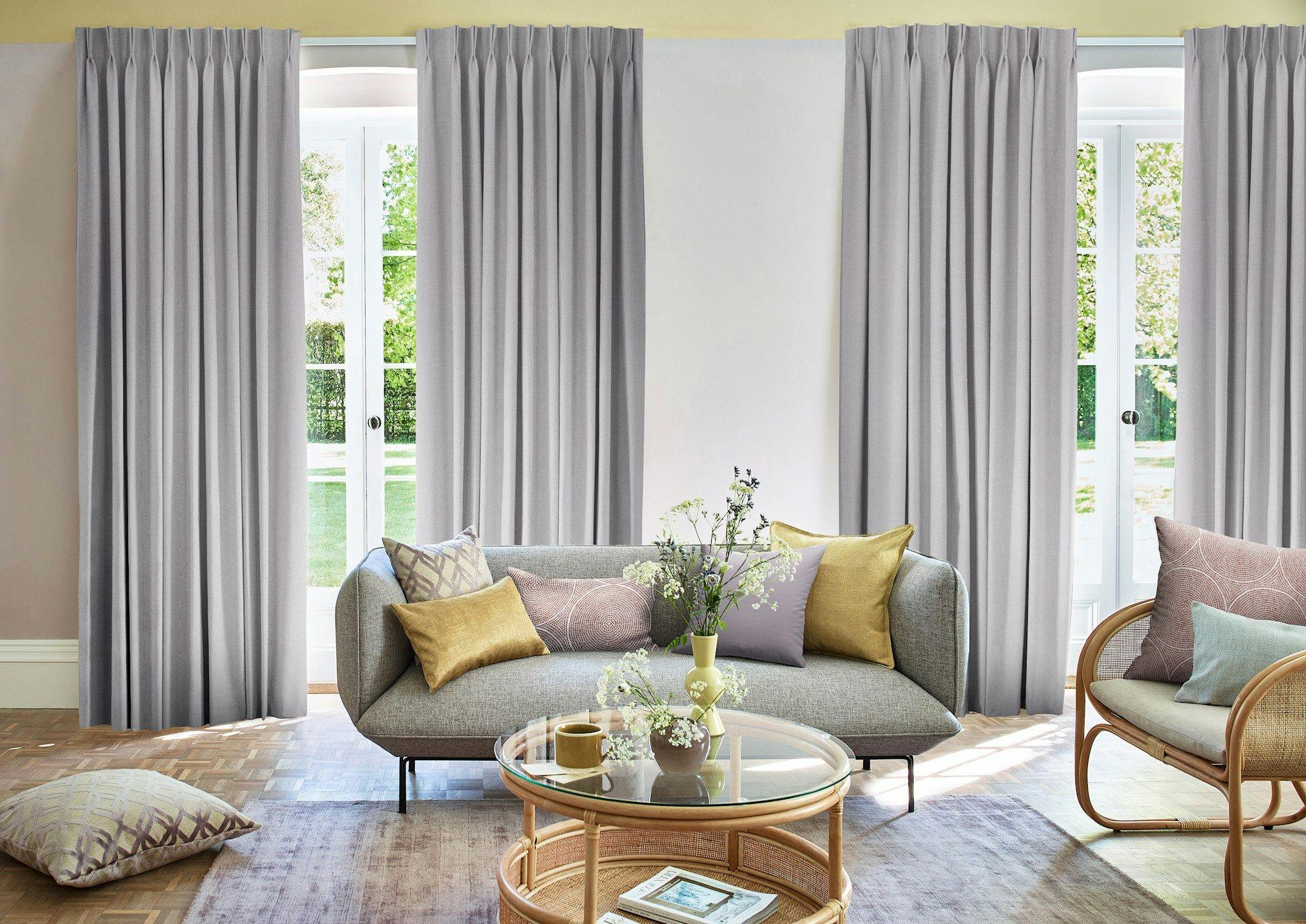 A contemporary living room features a gray sofa with assorted cushions, a round glass coffee table with flowers, and tall gray curtains framing large windows. Sunlight highlights the wooden floor.