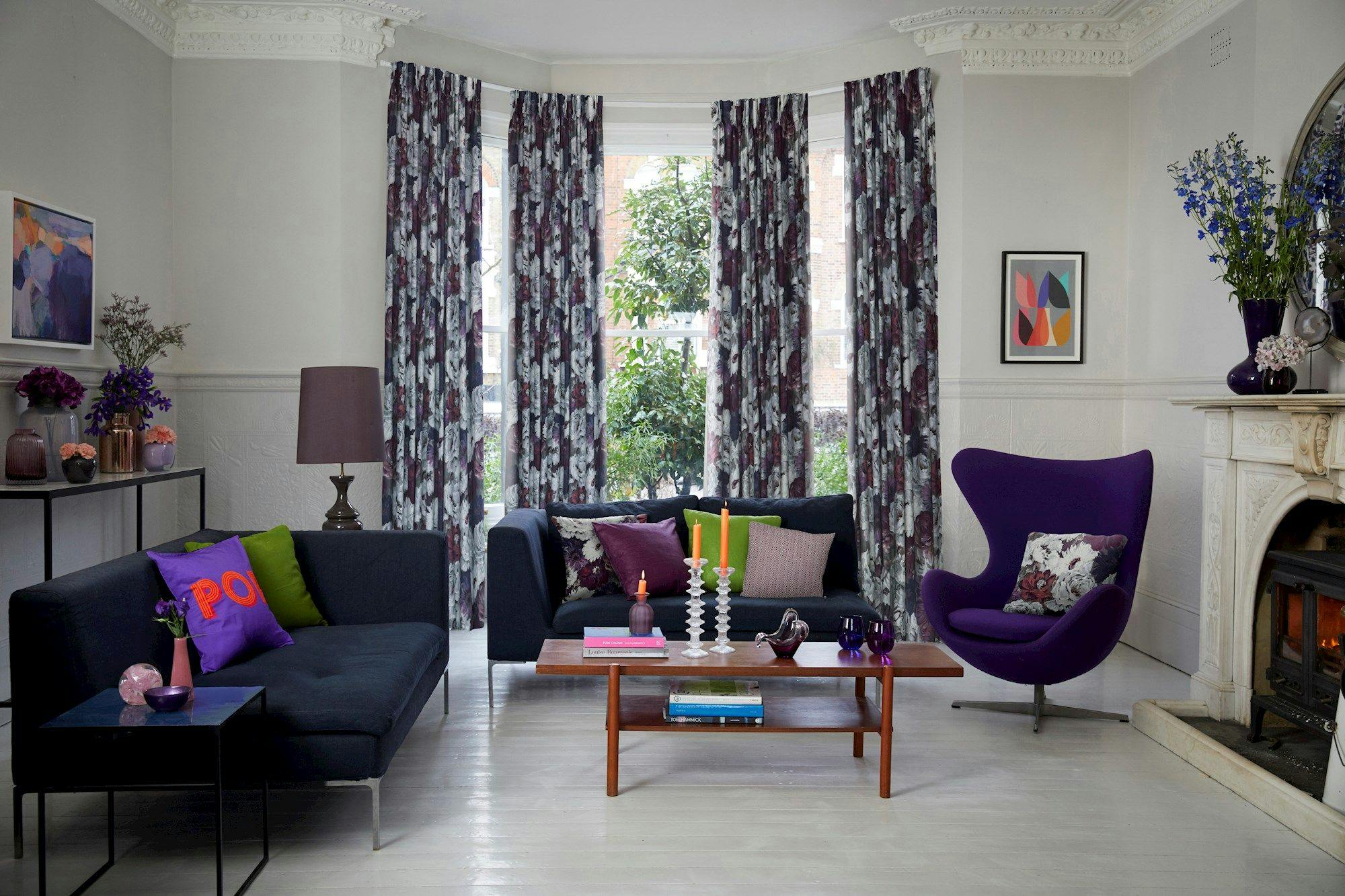 Sofa with colorful cushions sits beside a modern coffee table holding candles and books, surrounded by patterned curtains and eclectic decor in a bright, elegant living room.