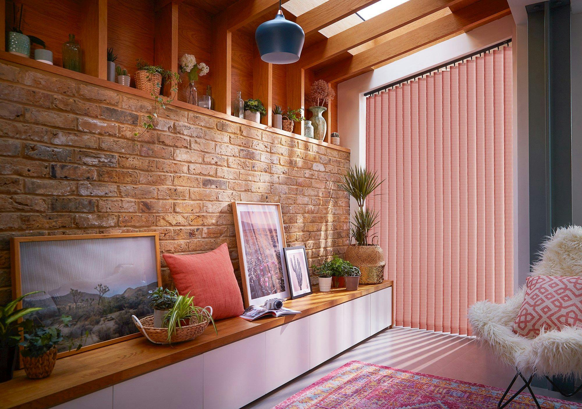 A cozy room features a long wooden bench displaying framed pictures, plants, and a coral pillow. Natural light filters through pink vertical blinds, highlighting a brick wall and decorative vases.