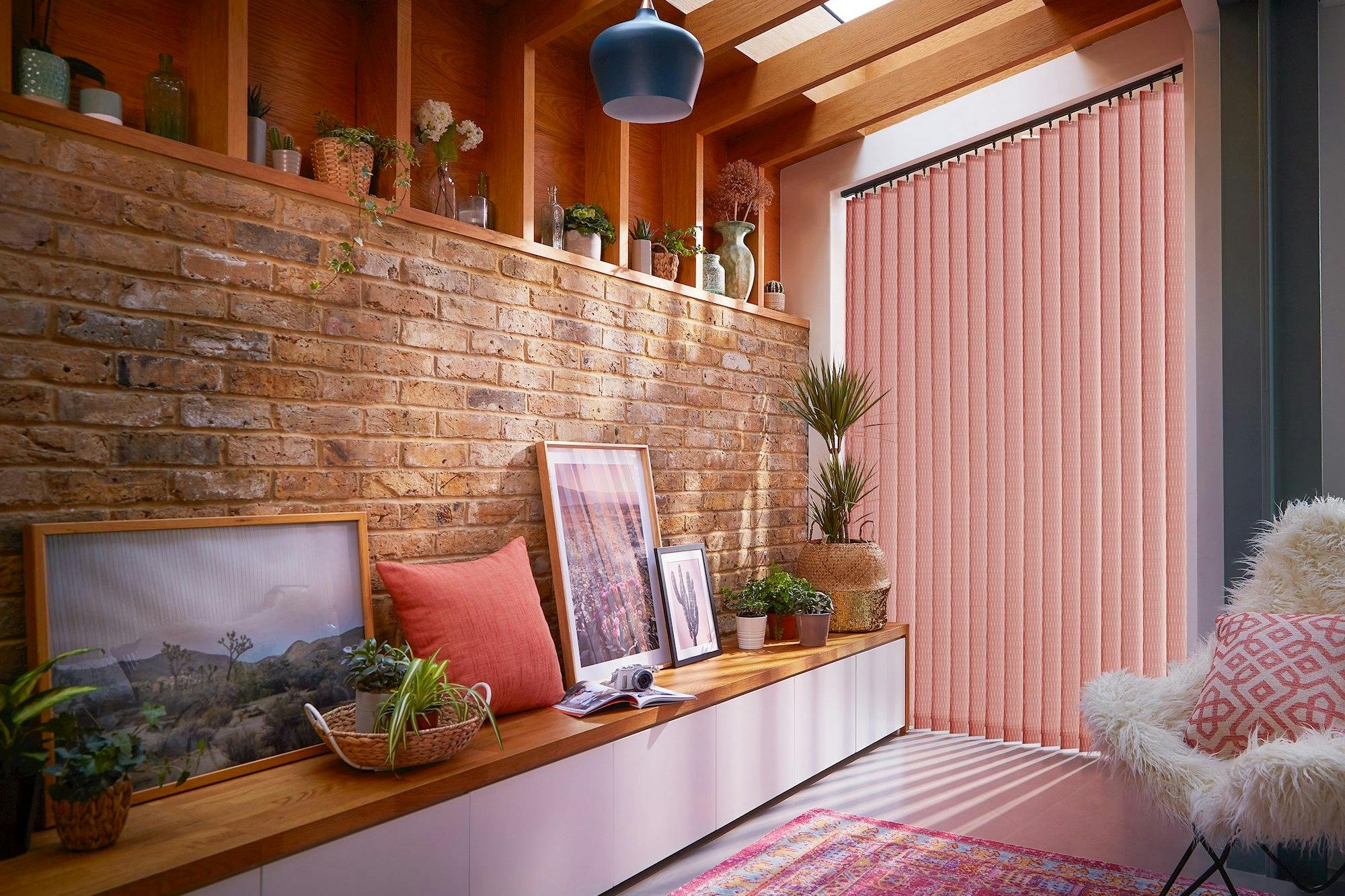 A cozy room features a long wooden bench displaying framed pictures, plants, and a coral pillow. Natural light filters through pink vertical blinds, highlighting a brick wall and decorative vases.