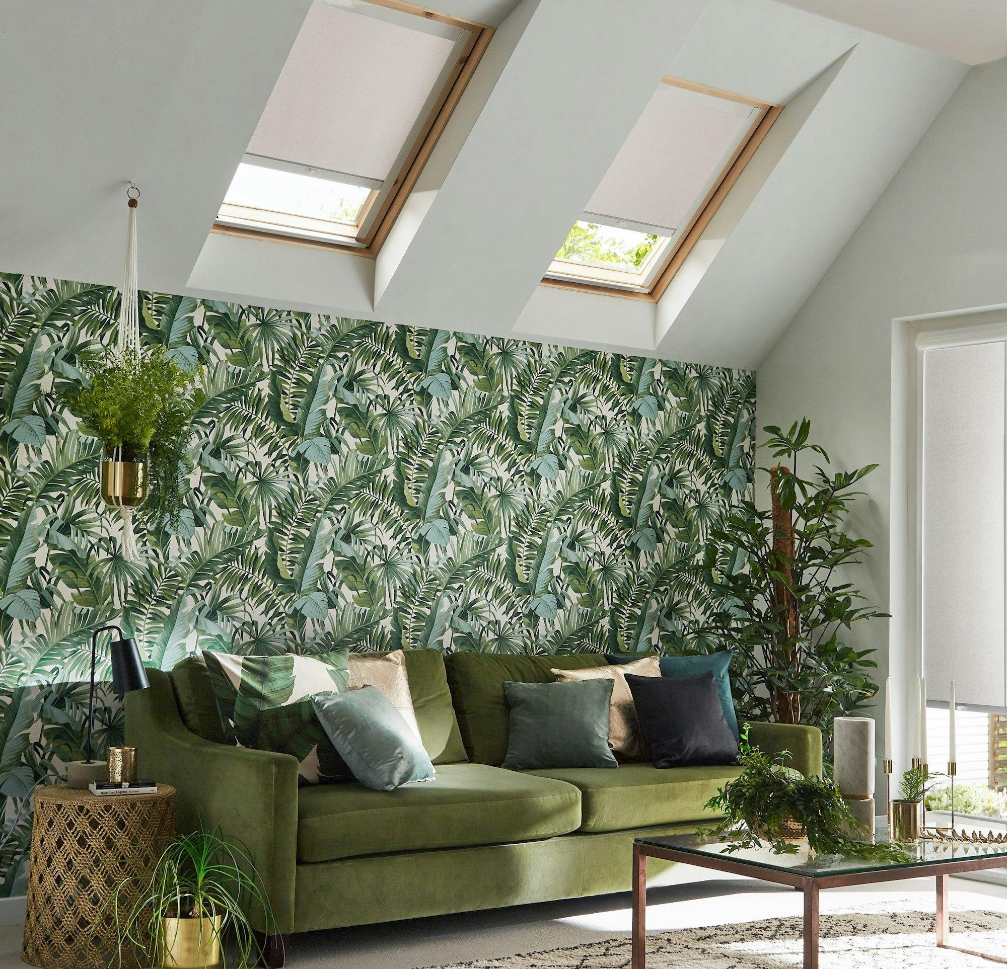 A green sofa adorned with multicolored cushions sits against a tropical leaf-patterned wallpaper in a sunlit room with skylights. Surrounding the sofa are plants and a glass coffee table.