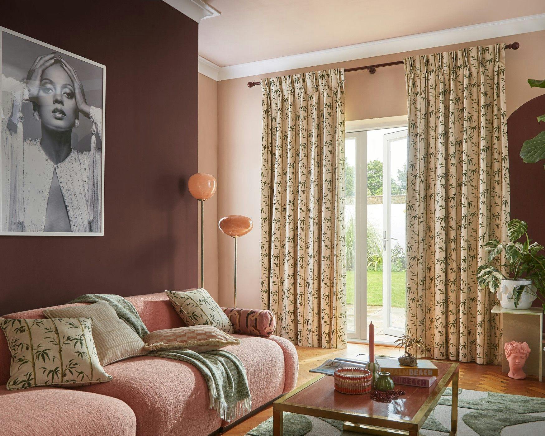 A pink sofa with patterned cushions sits in a cozy living room, featuring a large portrait on a dark wall. Curtains with leaf designs hang beside a glass door overlooking greenery.