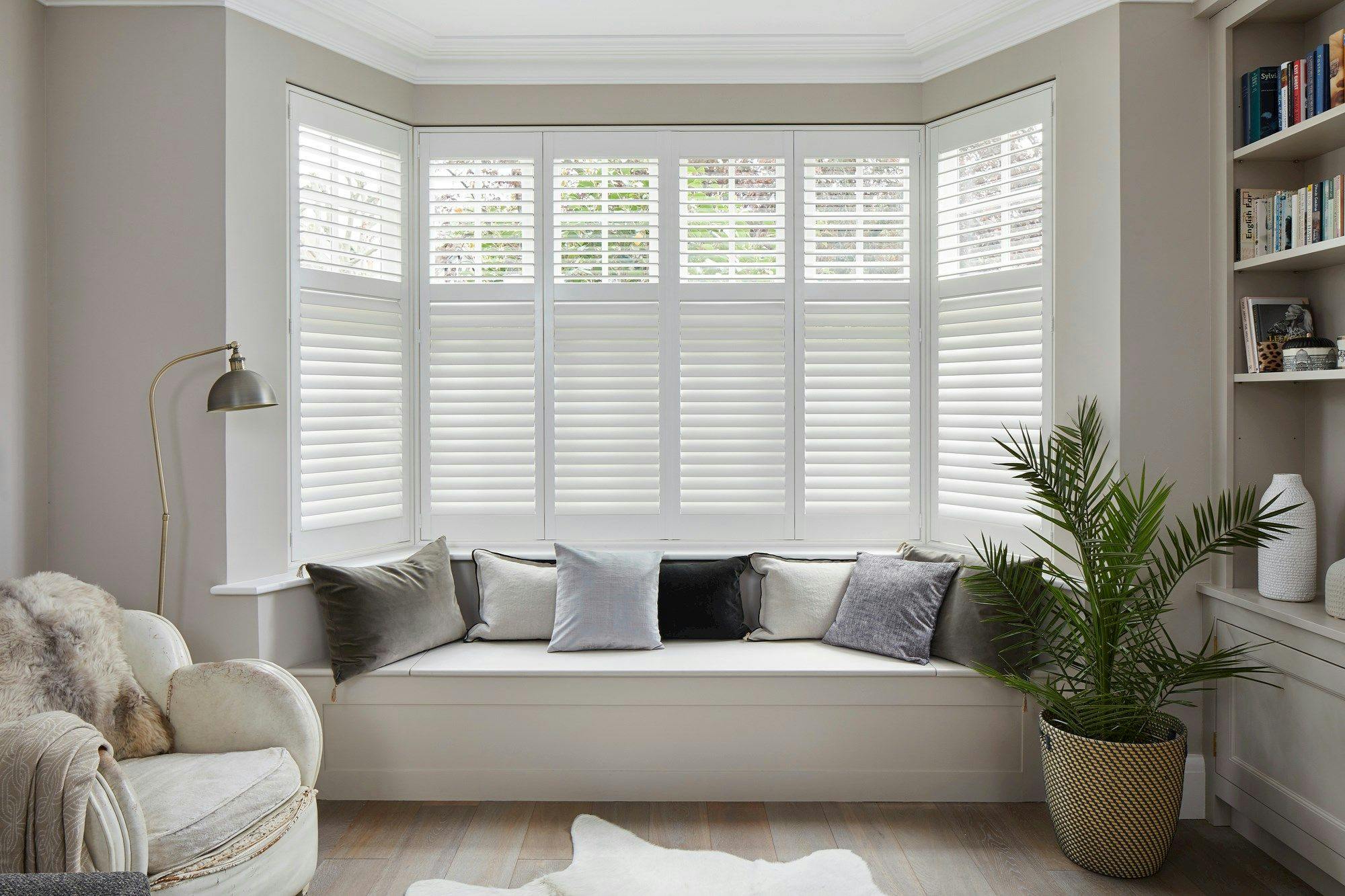 White shutters in a living room with a sofa facing away from the windows. A potted plant and bookshelf on the right hand side. 