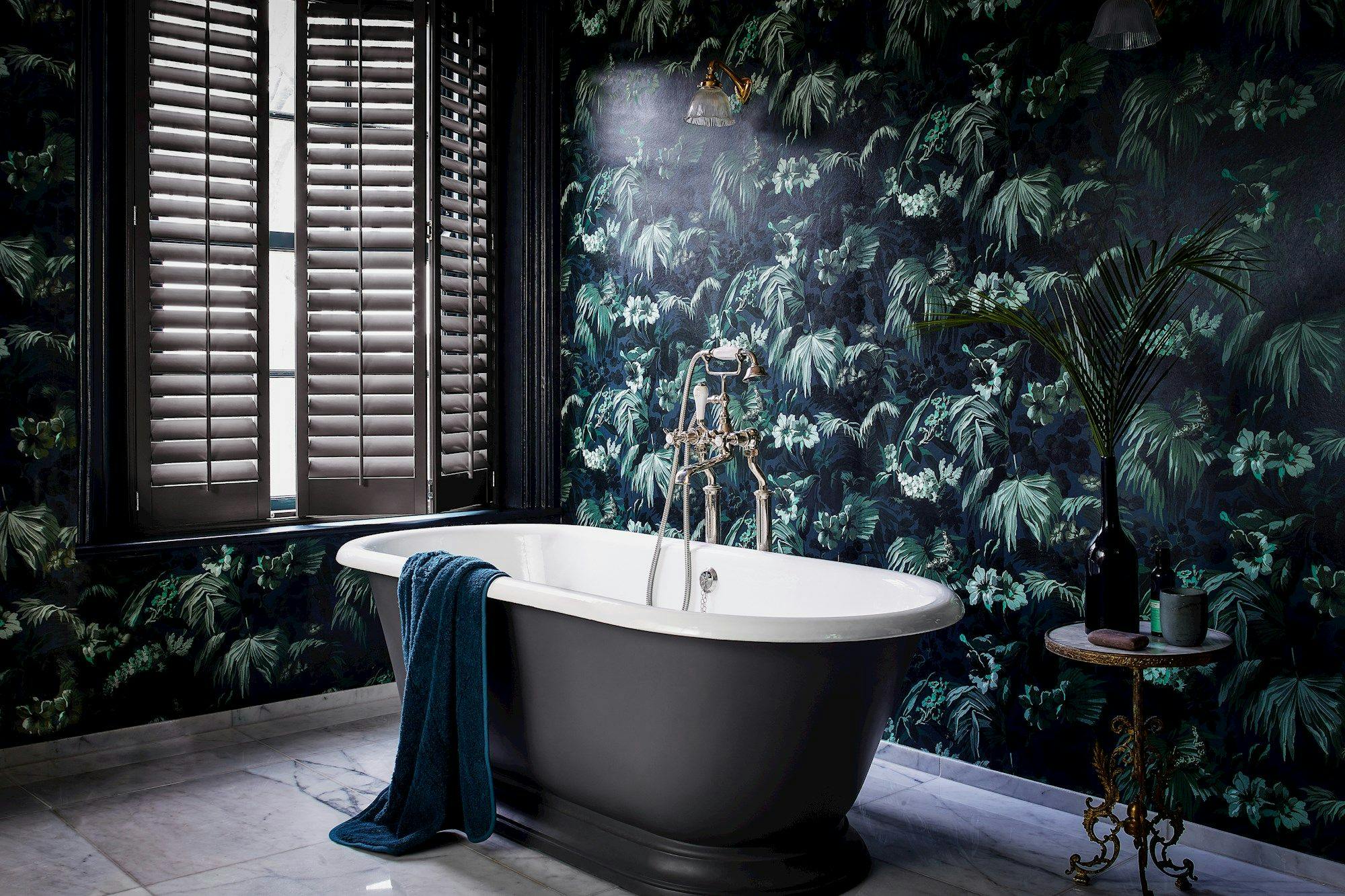 A freestanding bathtub with a blue towel drapes over the edge, sitting in a luxurious bathroom with tropical wallpaper and closed wooden shutters.