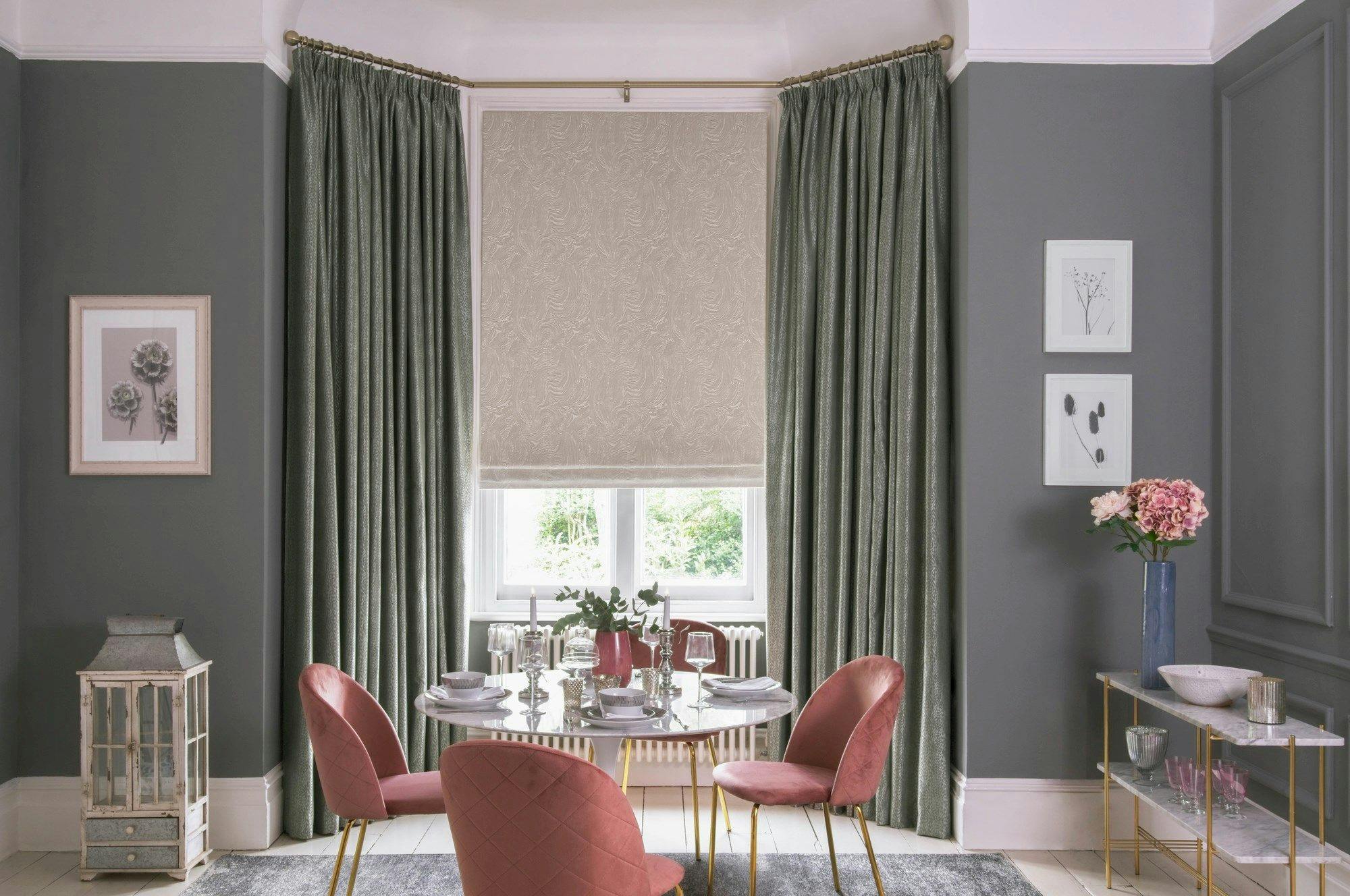 A round dining table set with dishes and glasses, surrounded by pink chairs, sits in a gray-walled room with green curtains and framed botanical art.