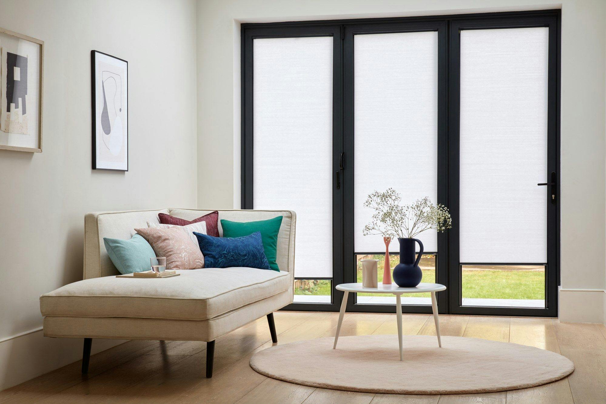 White Pleated blinds dressed on bi-fold doors of living room. Red, cream, green and blue cushions have been placed on cream corner sofa bed in room.