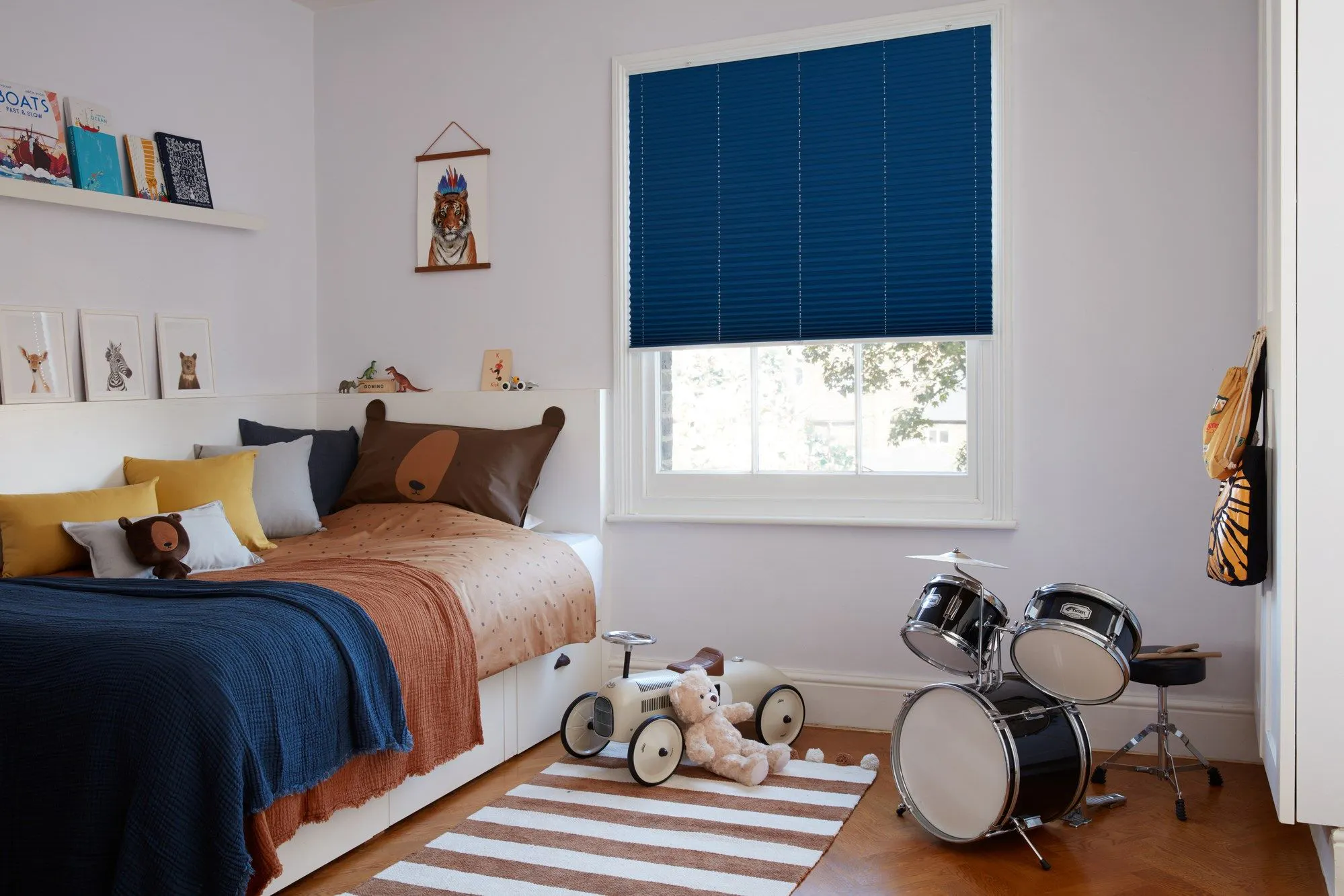 Dark blue Pleated blinds dressed on windows of kids' bedroom. Room has a single bed covered with brown cover, blue throws and cushions. A car, soft toy and drums are placed in the room.