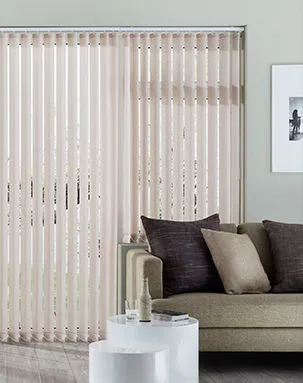 Vertical blinds cover a window, partially open to allow light. A beige sofa with brown and beige cushions sits nearby, accompanied by two round, white tables in a minimalist room.