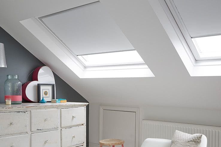 Two closed skylights illuminate a cozy attic room. A white dresser with decor items stands on the left, while a white couch and small round stool are nearby.