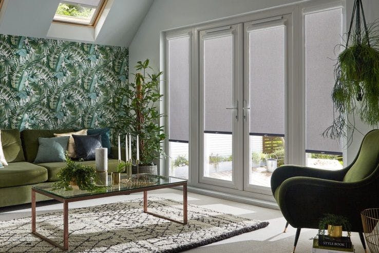 four light grey perfect fit blinds partially open on door length windows in a grey room with green furnishings and plants including a feature wall of plant themed wallpaper