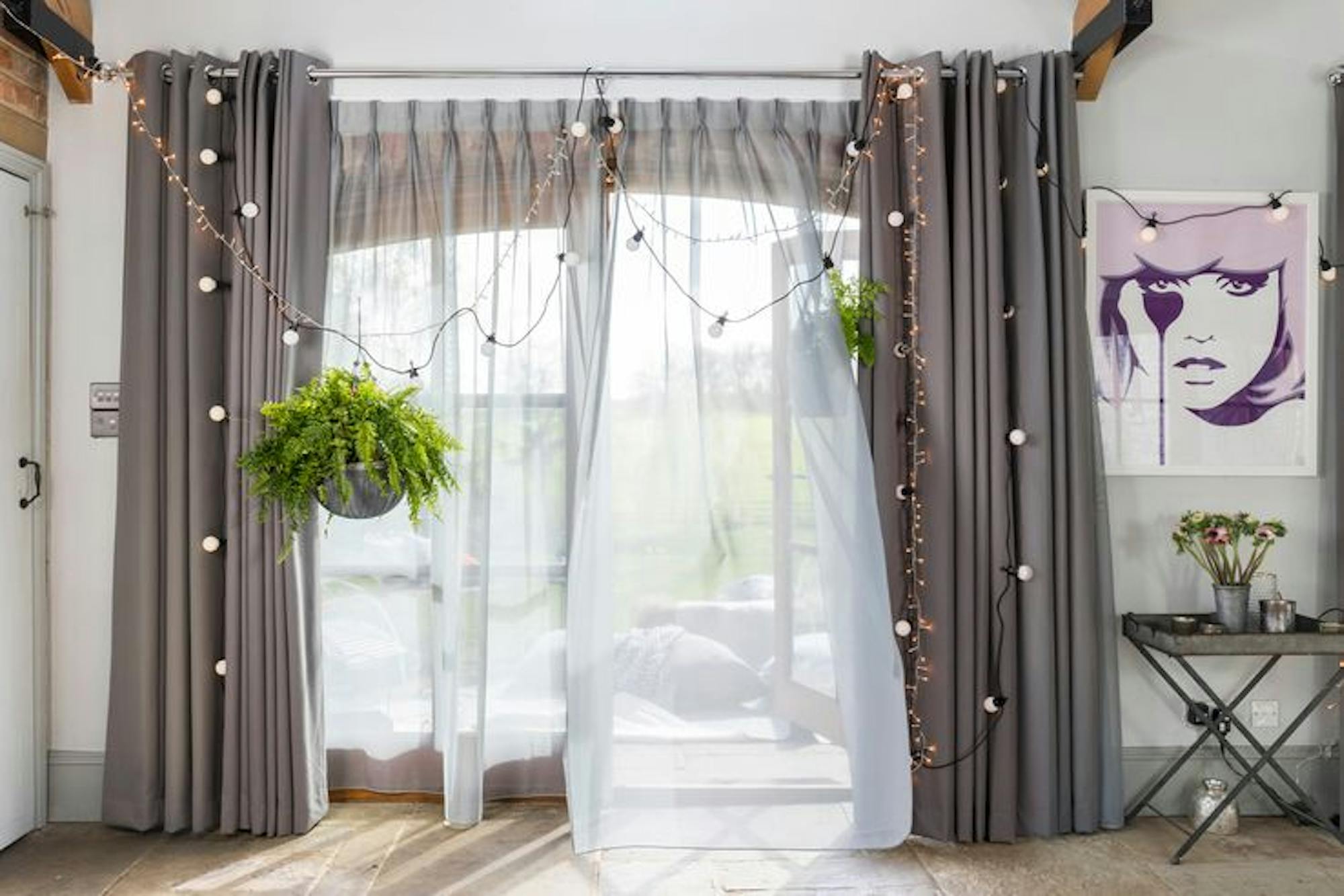 Curtains drape over sliding glass doors adorned with string lights and a hanging plant. Nearby, a poster of a stylized face hangs above a small table with potted plants.