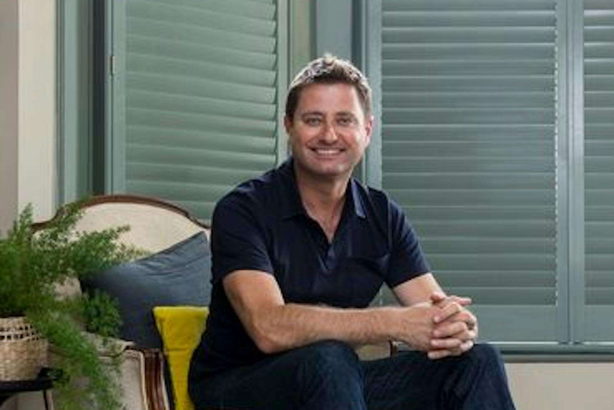 George clarke sat on a cream chair in a living room that has teal coloured shutters