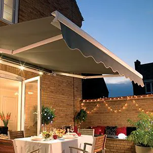An outdoor patio with a dining table set for a meal, beneath an extended awning. The brick-walled area is decorated with string lights and plants, creating a cozy evening atmosphere.
