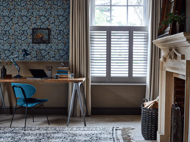 Home office with blue floral wallpaper window with pale grey cafe style shutters, partially open, dark cream curtains, wooden trestle style desk with blue chair, blue patterned rug on wood floor, wood fire surround.