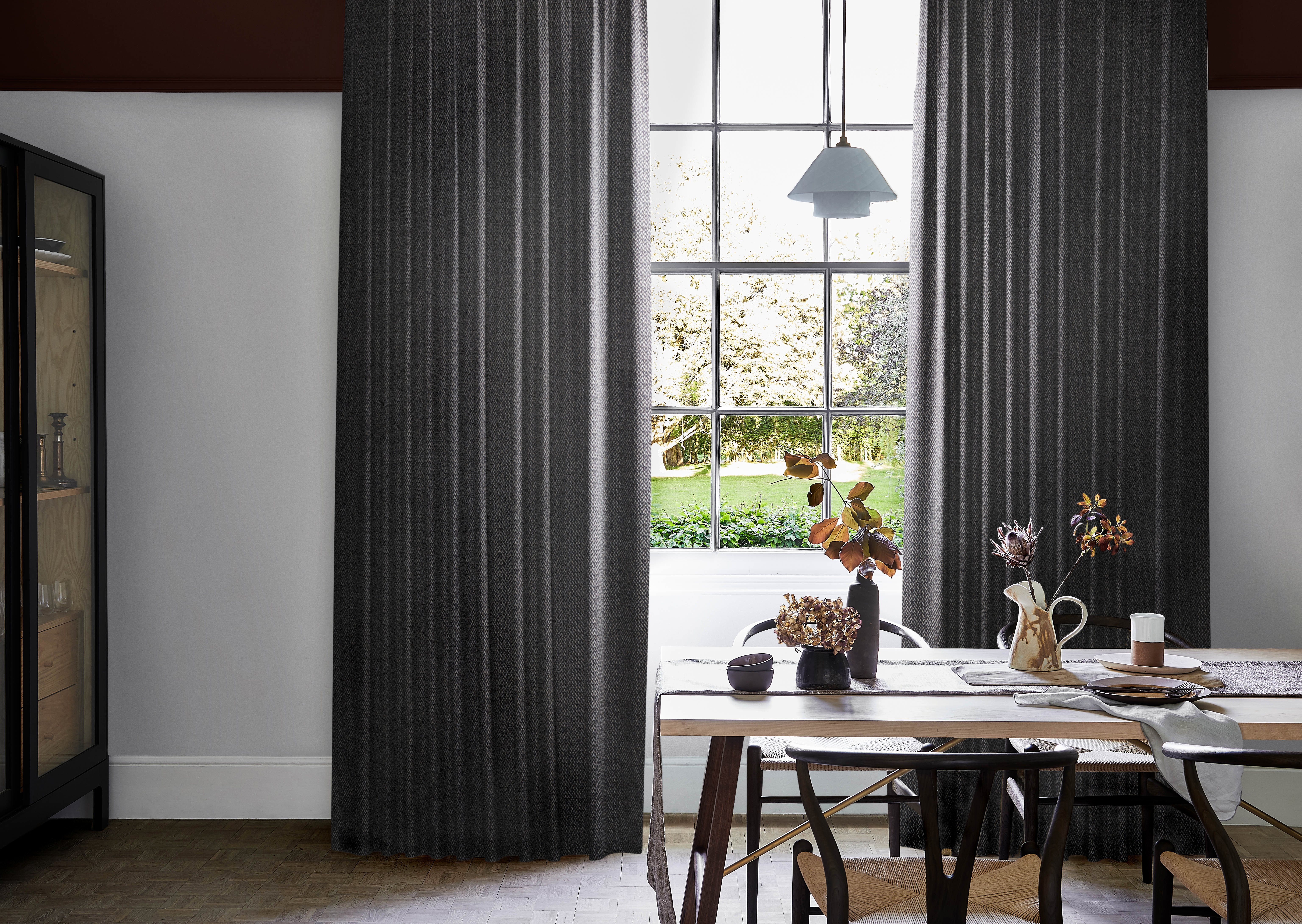 Modern dining area with dark graphite wave curtains framing large grid window, wooden table set with ceramic dishes, vases, and dried foliage.