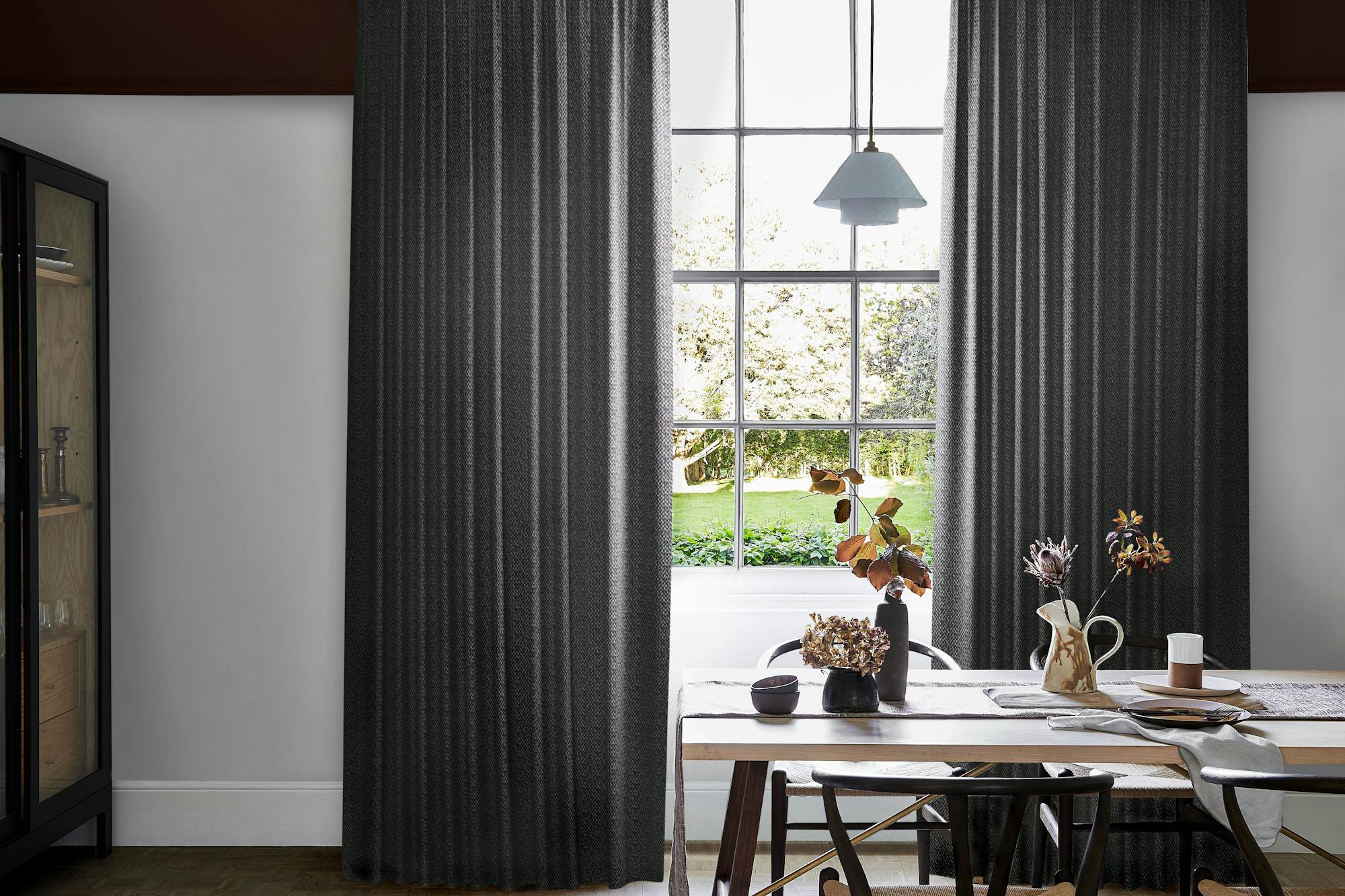 Modern dining area with dark graphite wave curtains framing large grid window, wooden table set with ceramic dishes, vases, and dried foliage.