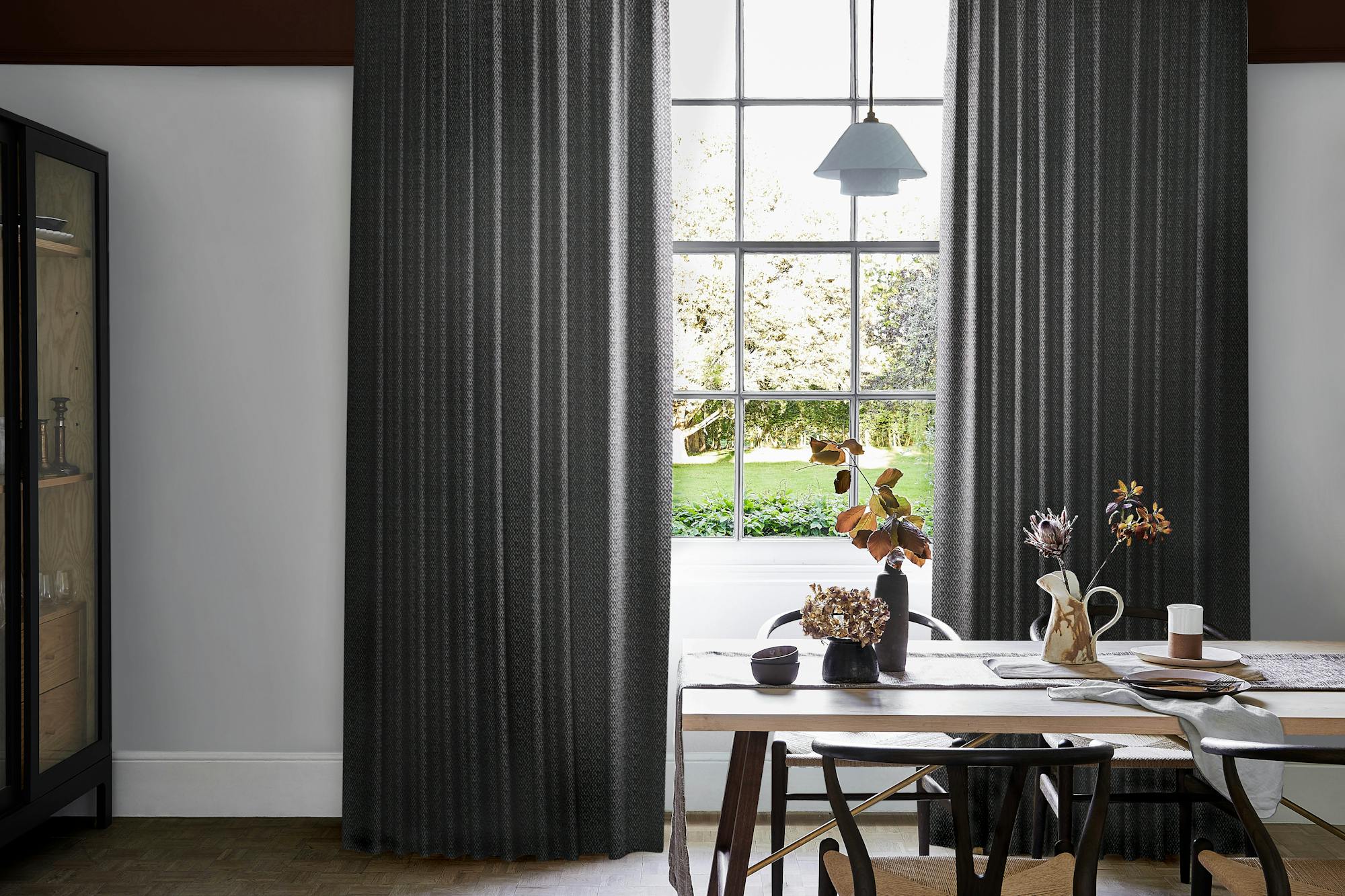 Modern dining area with dark graphite wave curtains framing large grid window, wooden table set with ceramic dishes, vases, and dried foliage.