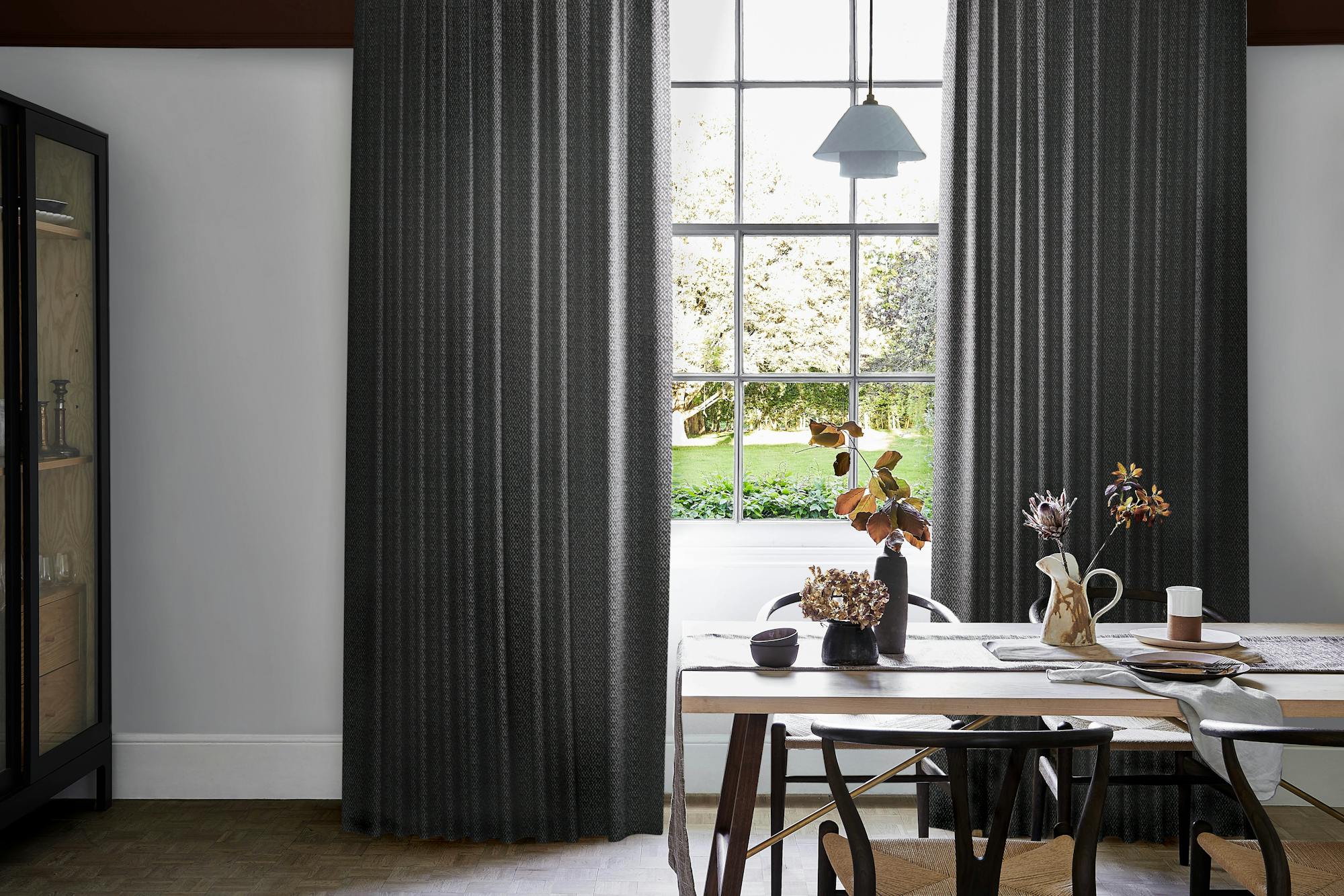 Modern dining area with dark graphite wave curtains framing large grid window, wooden table set with ceramic dishes, vases, and dried foliage.