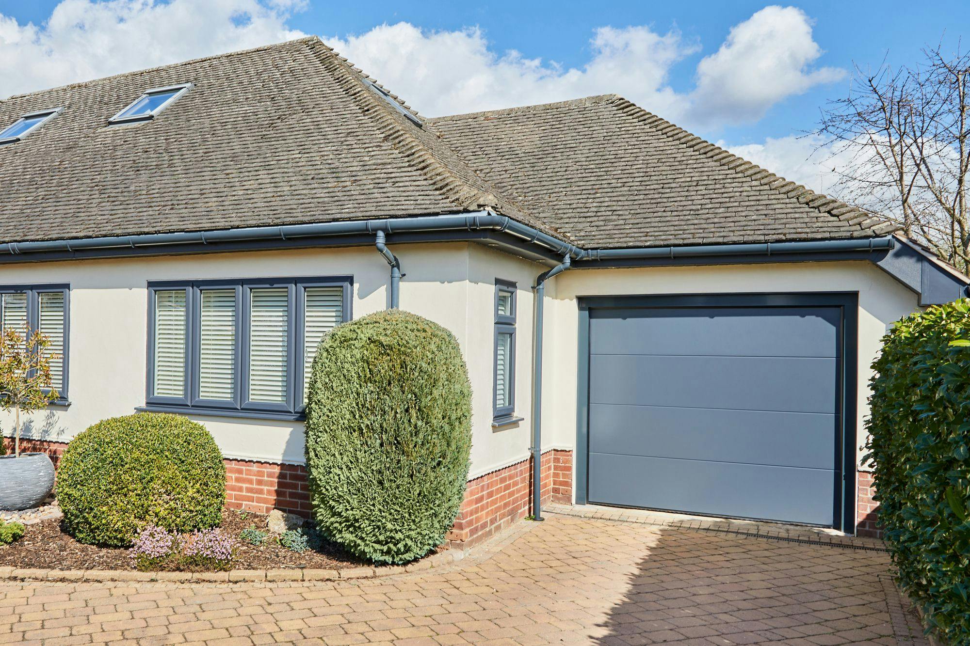 Corner shot of single storey house in white with anthracite grey integral sectional garage door matching guttering and window frames and block paved driveway.