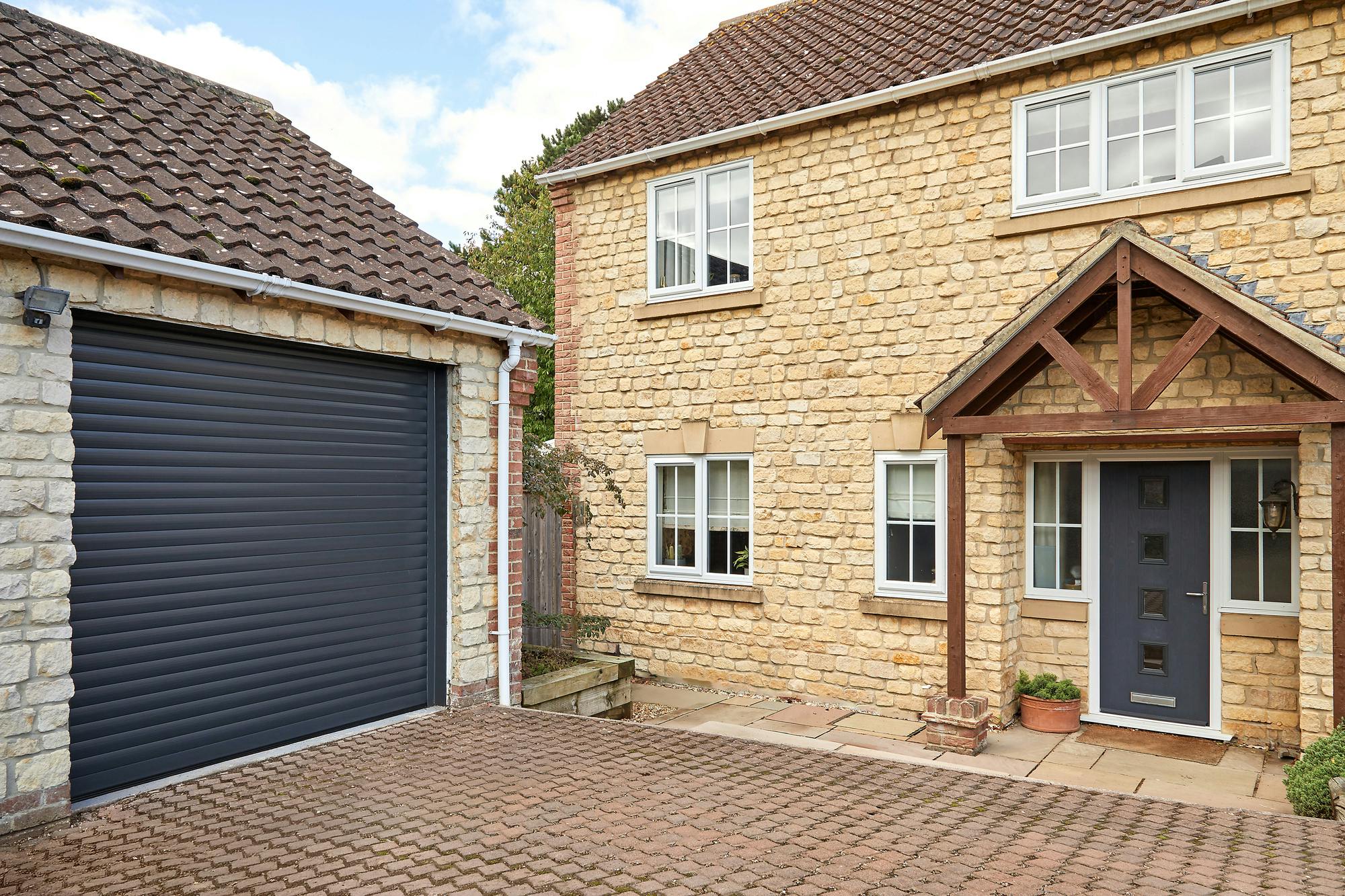 Traditional yellow-brown brick house with adjacent garage at 45 degree angle with an anthracite roller garage door, closed, a brick driveway connects the two, with a pergola porch over the house door which is a matching anthracite with four small square windows.