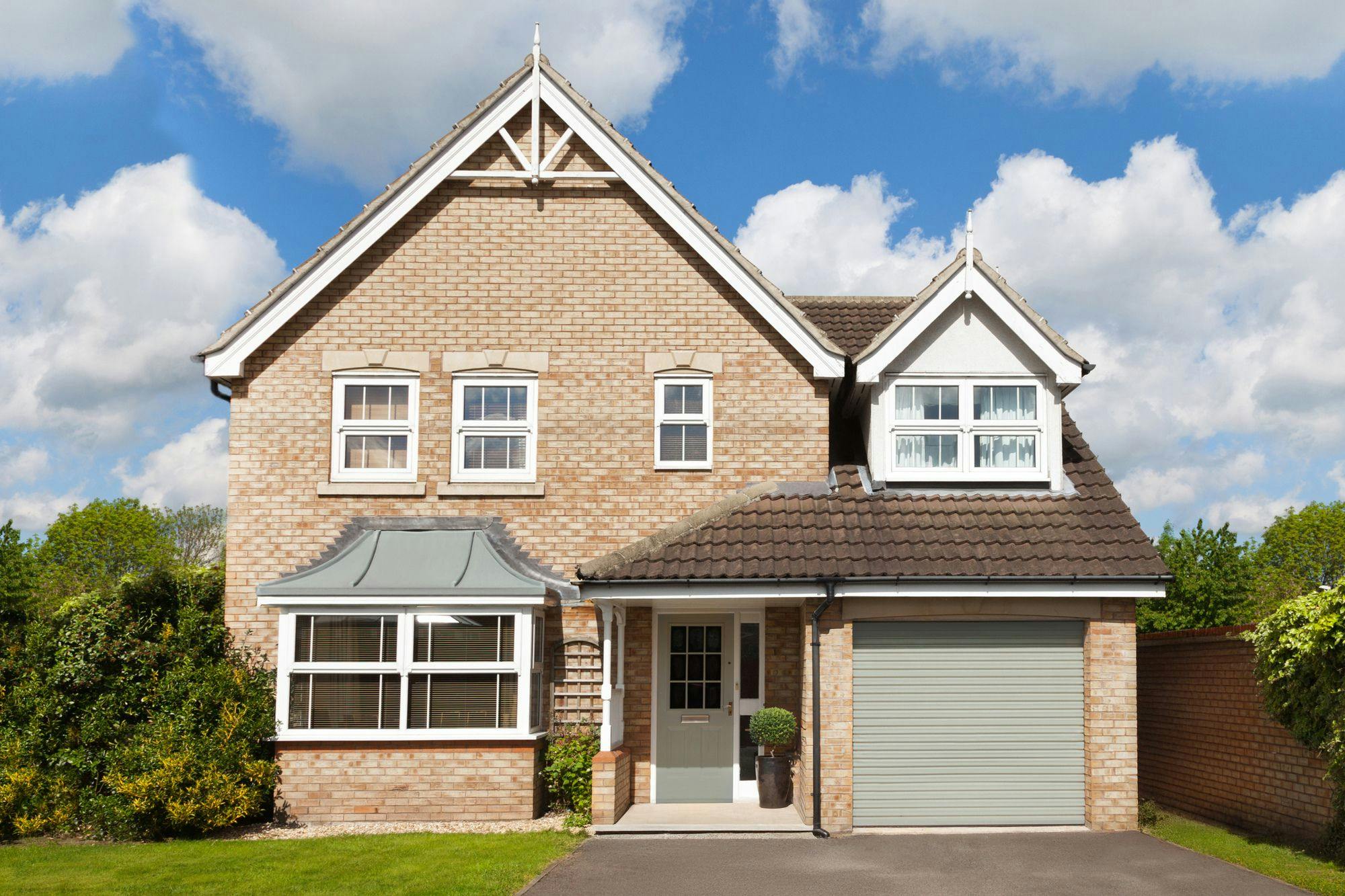 Exterior shot of large modern brown-brick house with integral garage door in pale grey-green, closed, white fascias to box bay window on ground floor, three small windows above bay and double window above garage, asphalt driveway with lawn to left and hedging.