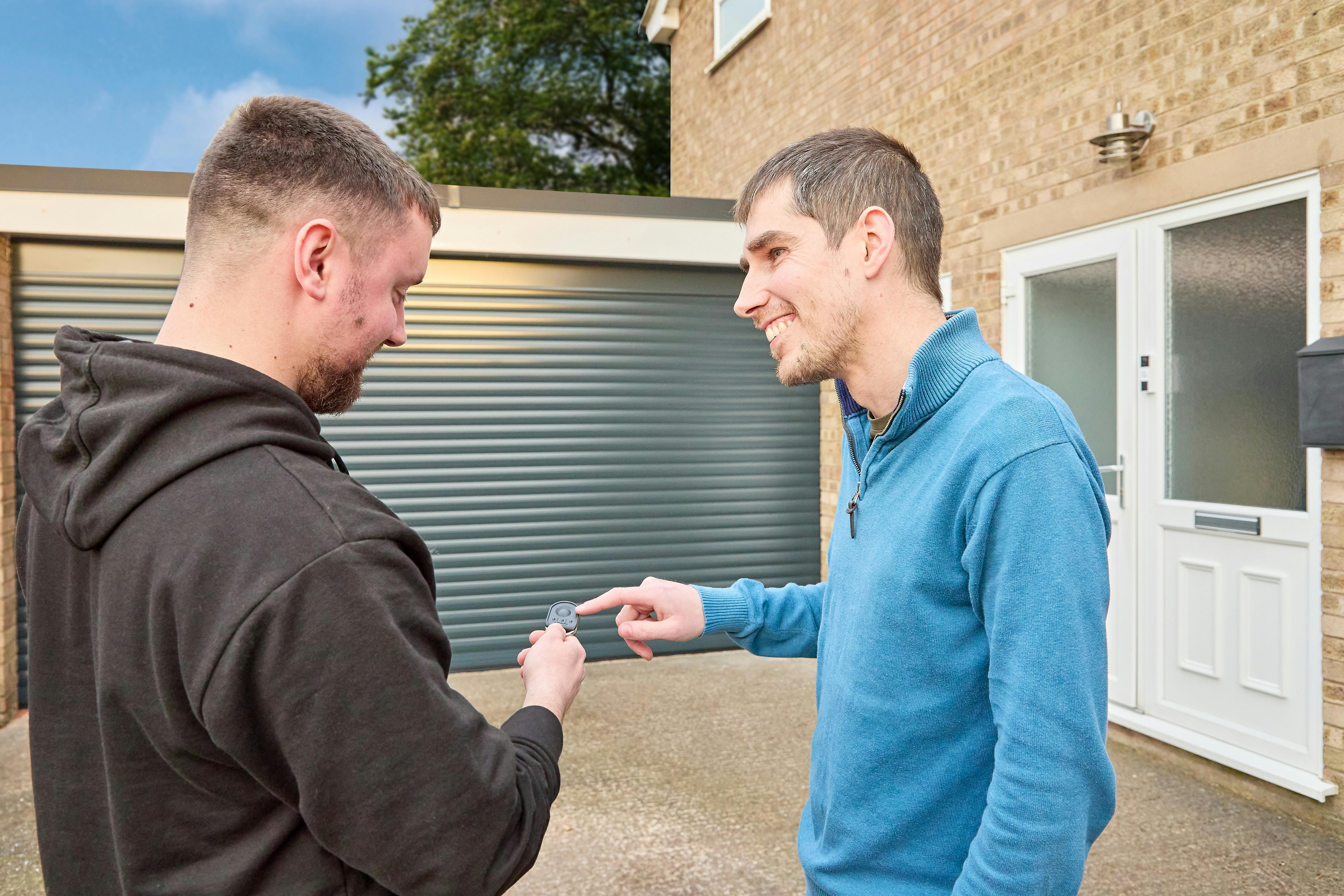 Hillarys advisor and male customer using a key fob pointing at a closed grey garage door on a concrete driveway outside a modern house