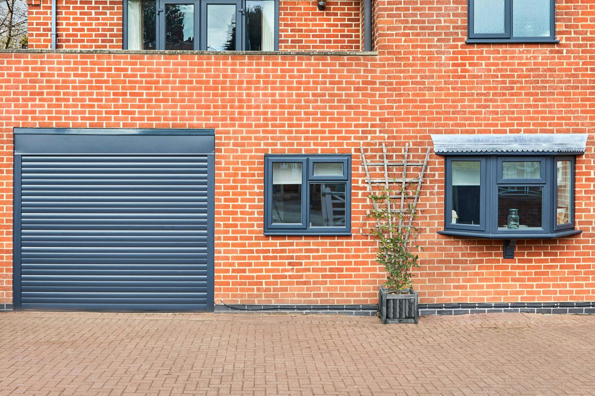 Large red brick modern house with integral garage, with anthracite grey door, matching window frames and pipes, matching front door,, dark wooden planter with climber and brick driveway across the whole front of the house.