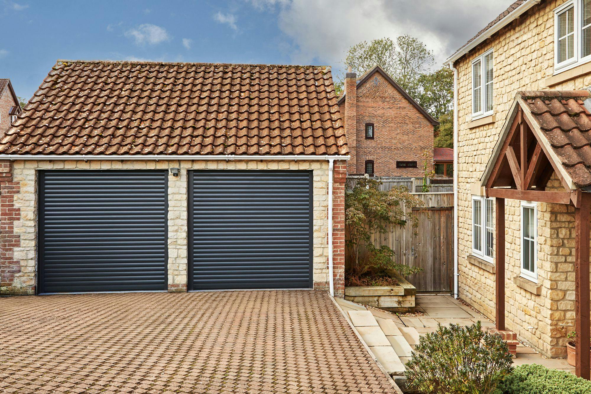 Traditional yellow-brown brick house with adjacent double garage at 45 degree angle with anthracite roller garage doors, closed, a brick driveway connects the two, with a pergola porch over the house door which is a matching anthracite with four small square windows.