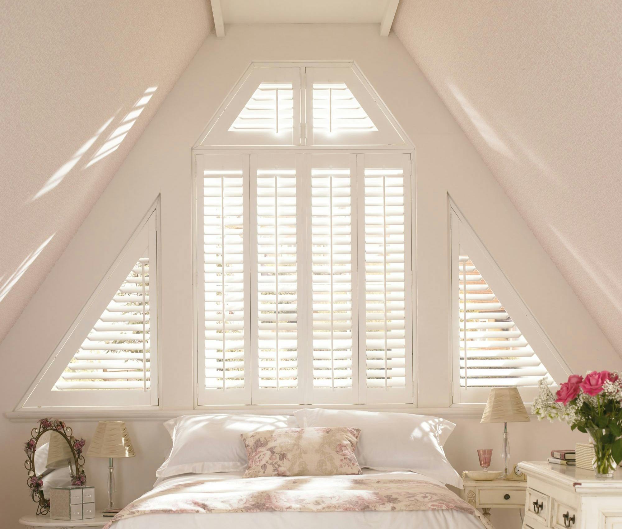 Cozy attic bedroom with white shutters, floral bedding, and vintage-style furniture in soft, natural light.