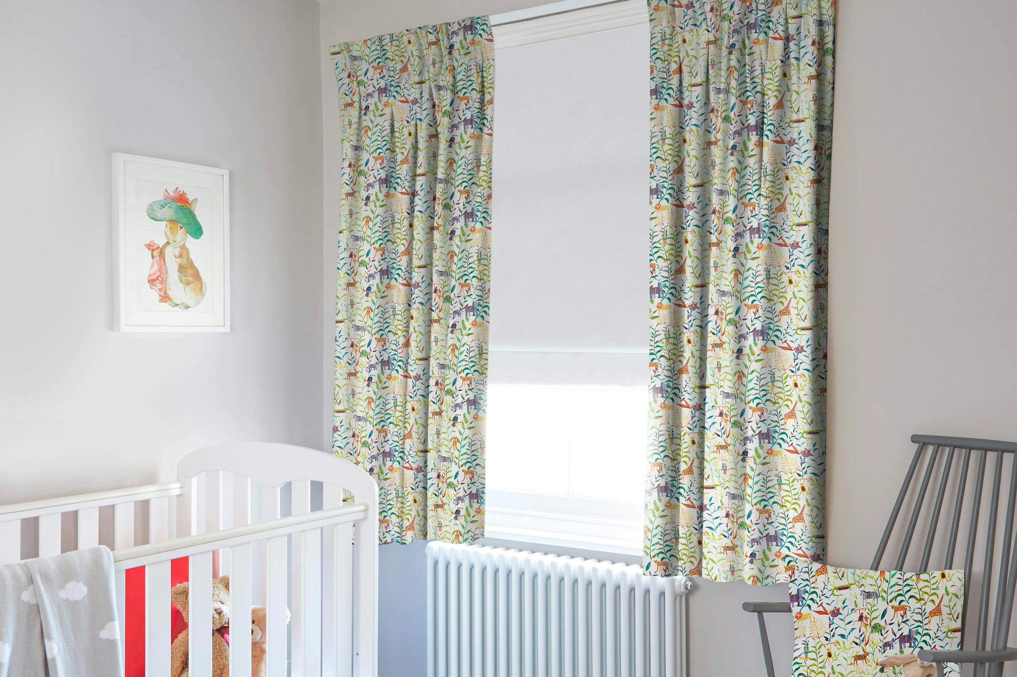 Child's bedroom in pale grey with window dressed in chalk white roller blind, lowered halfway under curtains in white with jungle animals in teal, umbre and ochre, grey wooden chair with cushion matching curtains, white cot with grey throw over. 