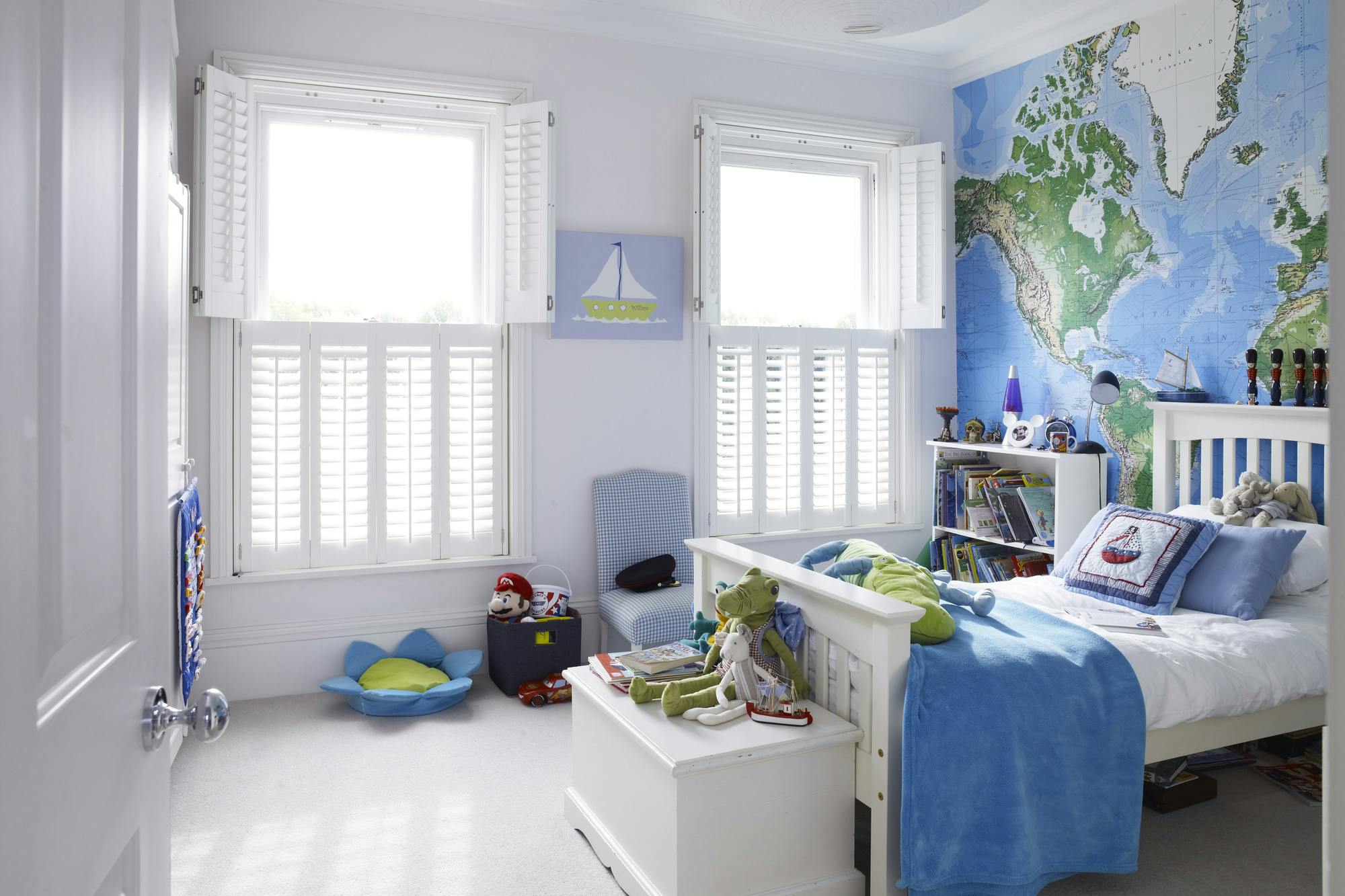 Child's bedroom in white with two adjacent windows dressed in white tier on tier shutters, top set open on both, white bed, feature wall behind map of the world, white bedding with blue throw and pillows, white toy box, white bookshelf, picture of sailboat between windows, white floor, white round paper lampshade to ceiling light. 