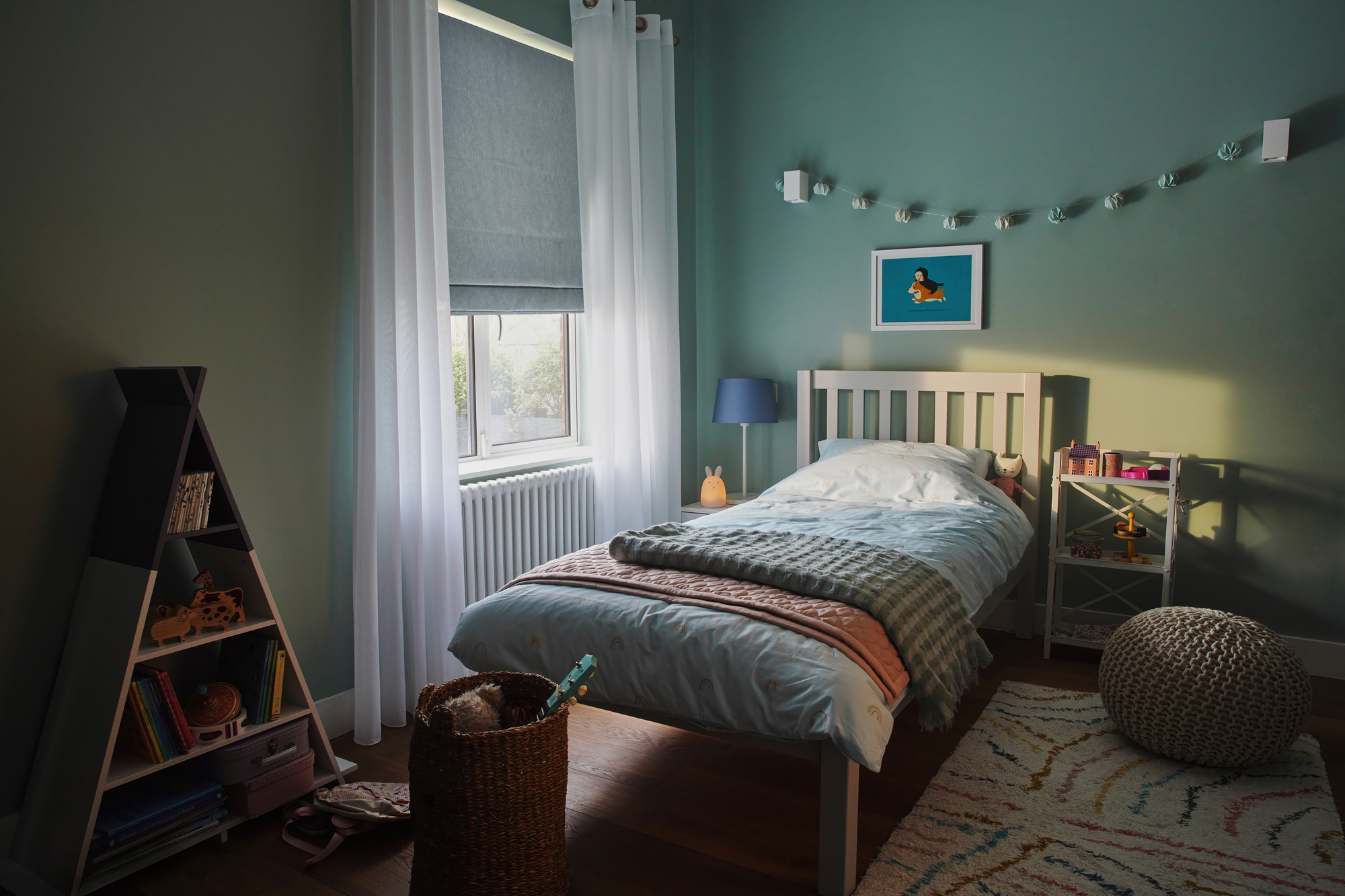 Child's bedroom in pale green-blue with window dressed in pale blue roman blind, lowered halfway and white sheer curtains, open, wooden bed with pale blue bedding with dark blue striped throw, blue bunting over bed, blue bedside lamp, triangular shelving unit with books and toys.