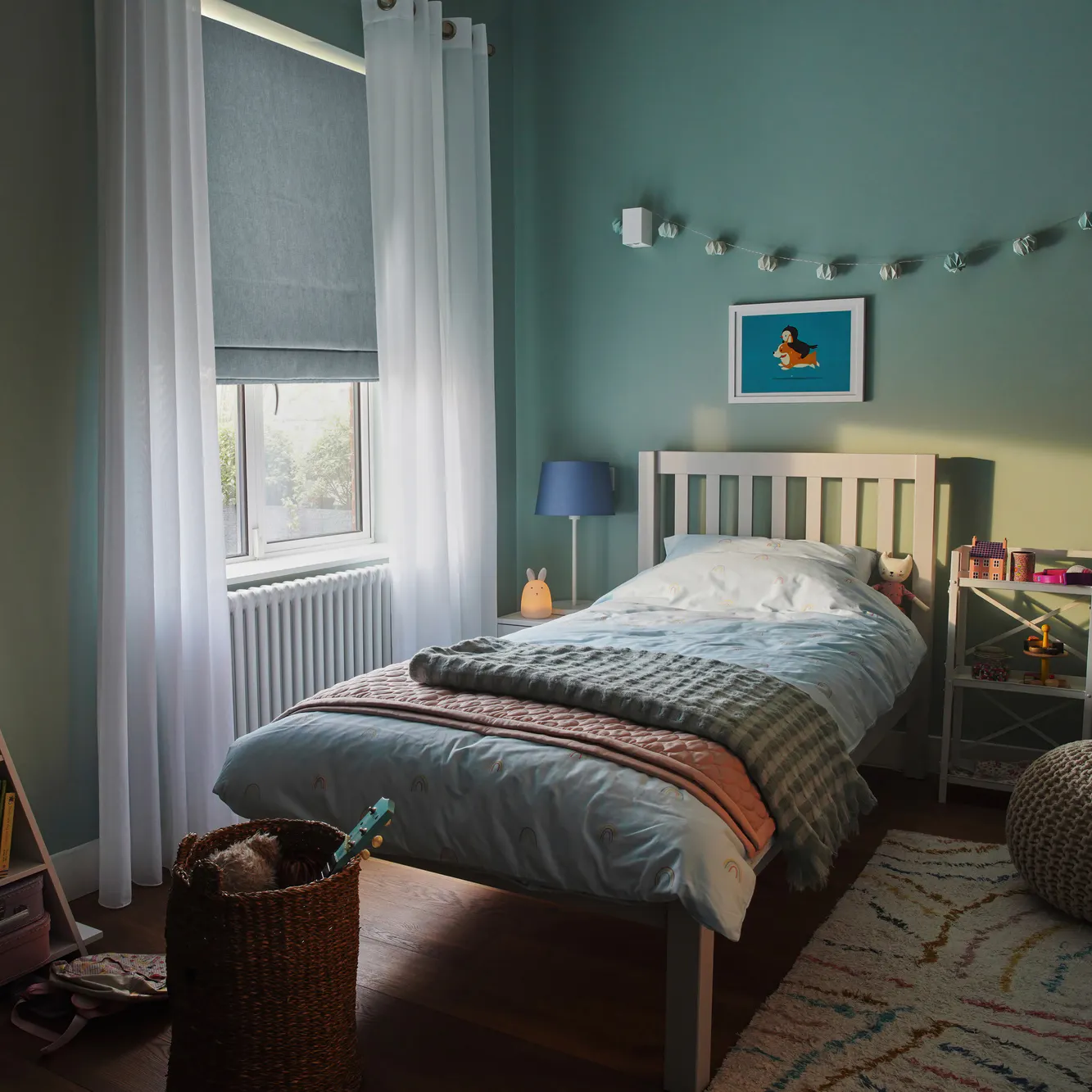Child's bedroom in pale green-blue with window dressed in pale blue roman blind, lowered halfway and white sheer curtains, open, wooden bed with pale blue bedding with dark blue striped throw, blue bunting over bed, blue bedside lamp, triangular shelving unit with books and toys.