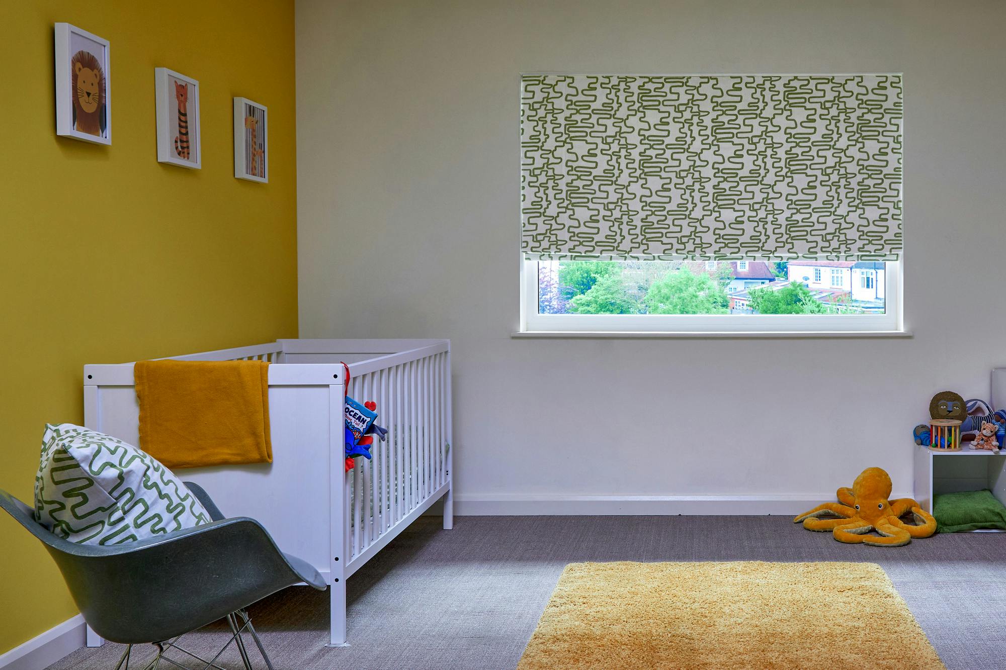 Child's bedroom in white with left hand wall mustard, window to rear dressed in white roller blind with black vertical scribble wave pattern, three quarters lowered, white cot, grey carpet with mustard rug in centre, modern rocker with cushion matching blind, mustard unit, toys around right hand side of room.