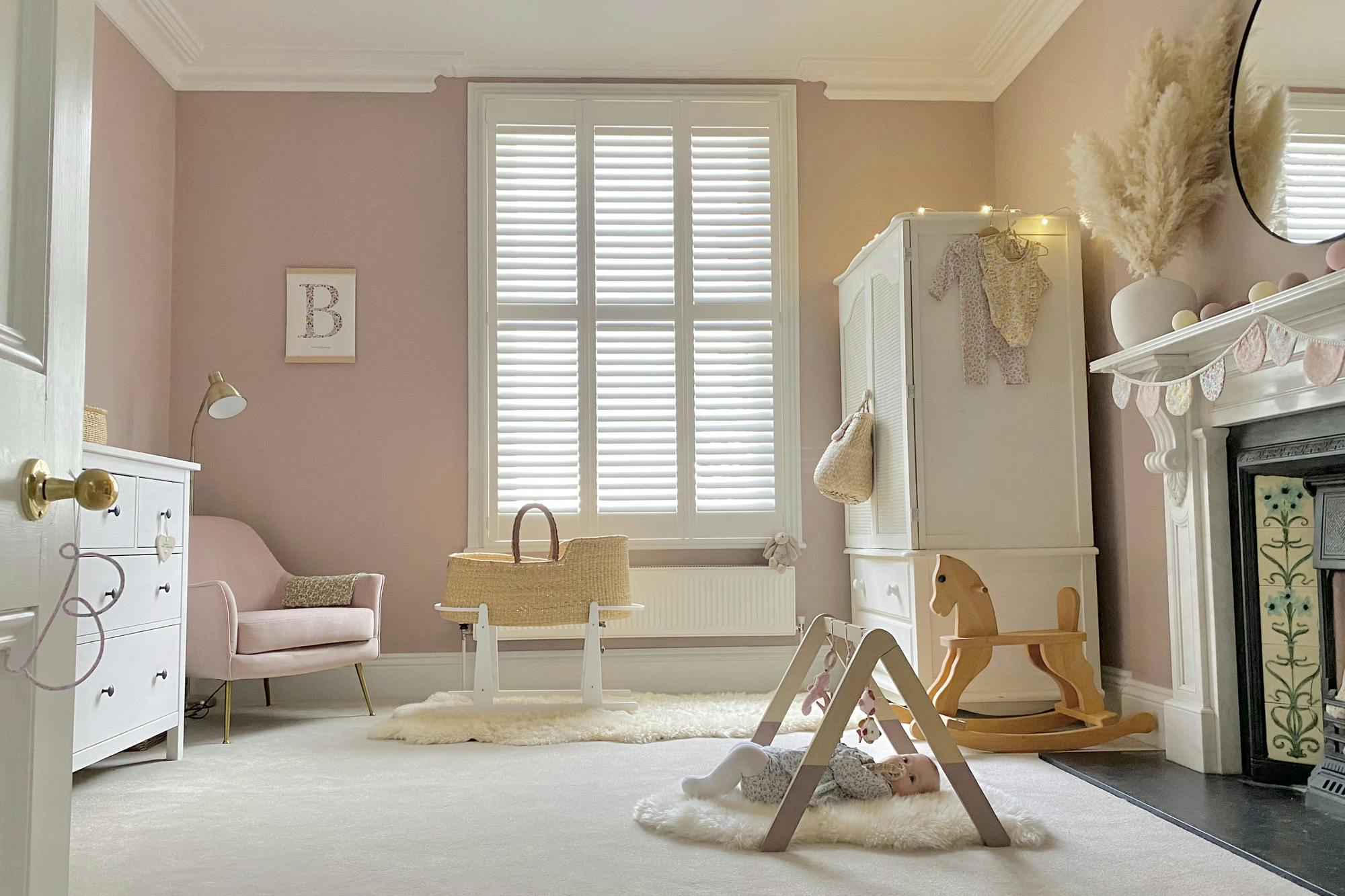 Baby's bedroom in soft pink with tall window dressed in white shutter, closed with louvres open, white wardrobe, moses basket on white stand on a cream sheepskin rug, pink chair, white chest of drawers, wooden rocking horse, white vase on white mantelpiece with pampas, pink and pale grey bunting, baby on sheepskin rug on cream carpet, under a wooden hanging toyframe.